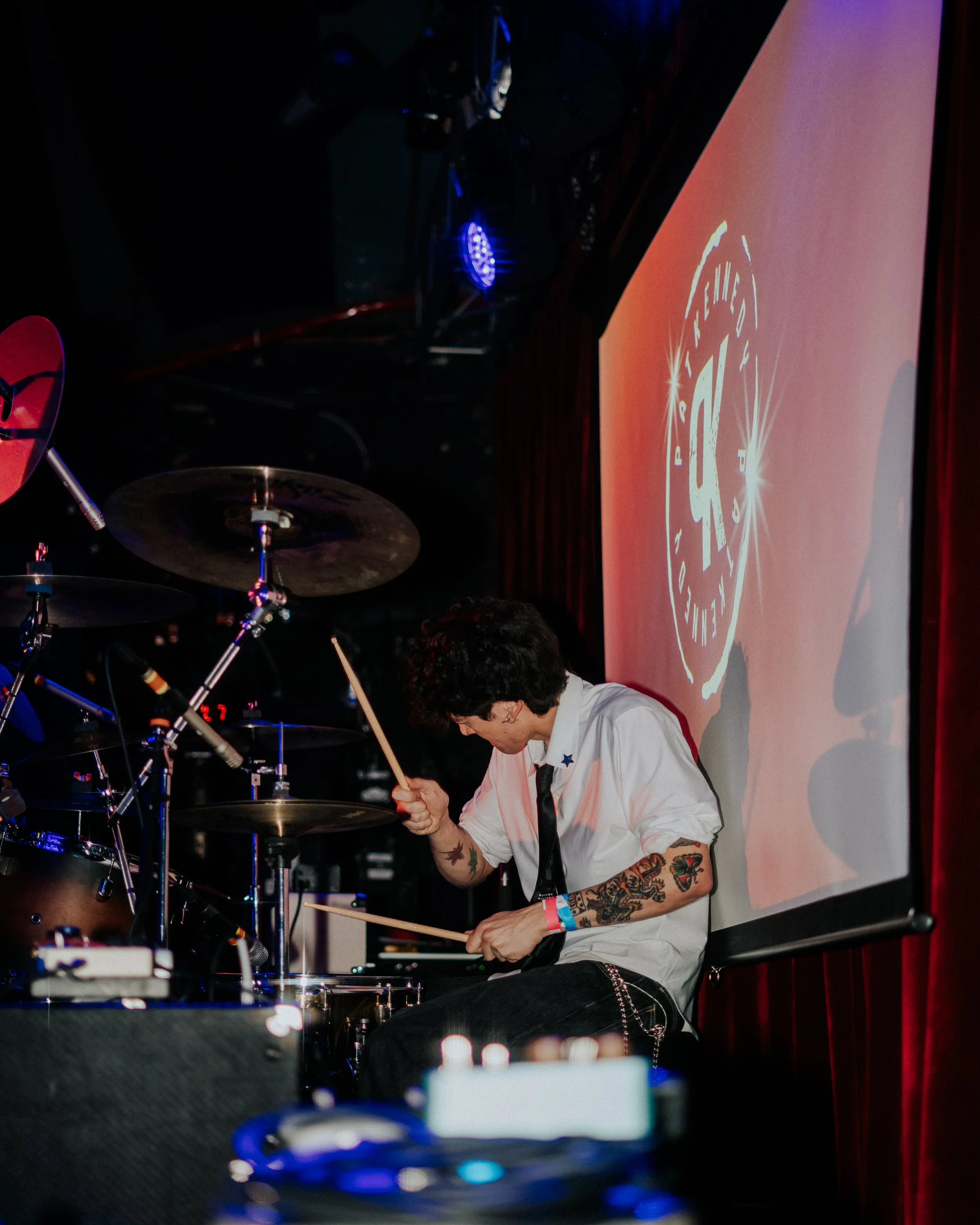 A person with short dark hair, wearing a white shirt with rolled-up sleeves, playing drum set on stage. The person has tattoos on their arms and wears a colored wristband. A large screen in the background displays a circular logo with text.