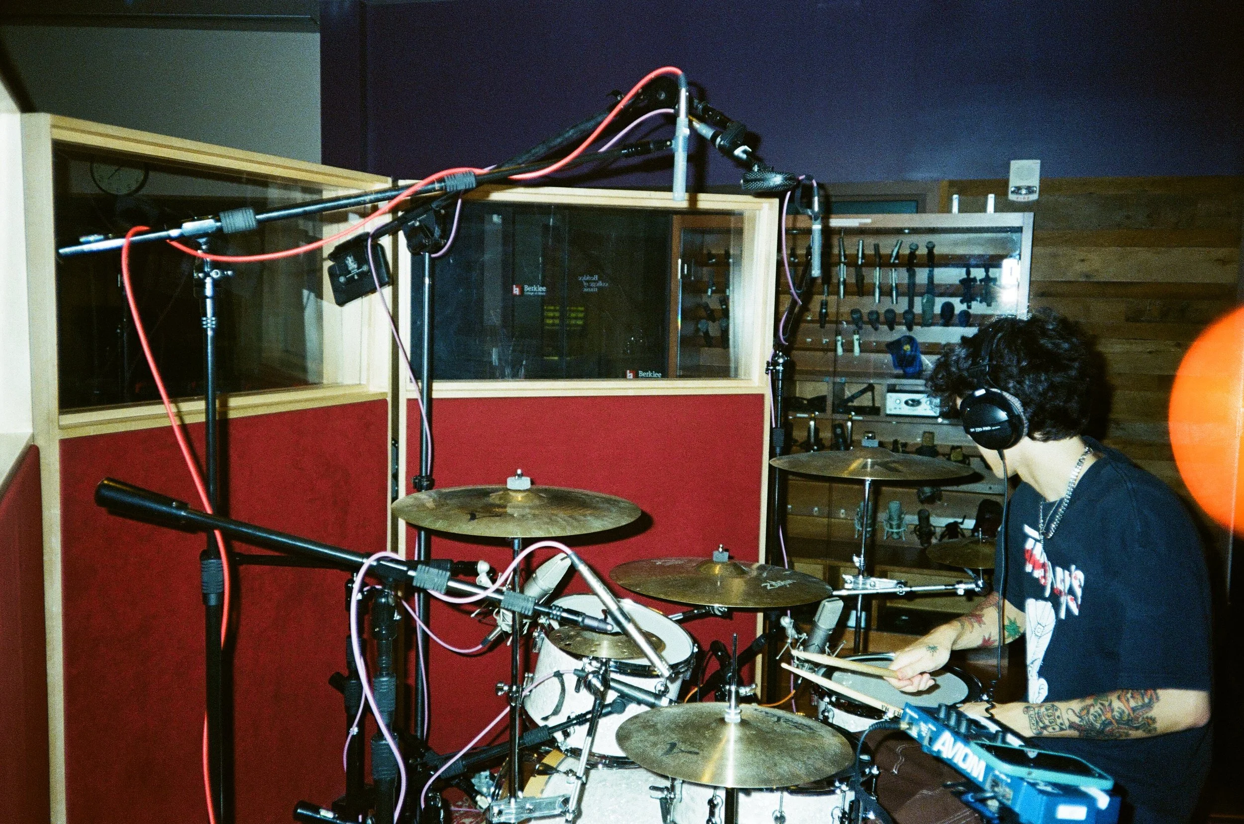 A person with tattoos playing a drum set in a recording studio with soundproofing walls, musical instruments, and recording equipment.