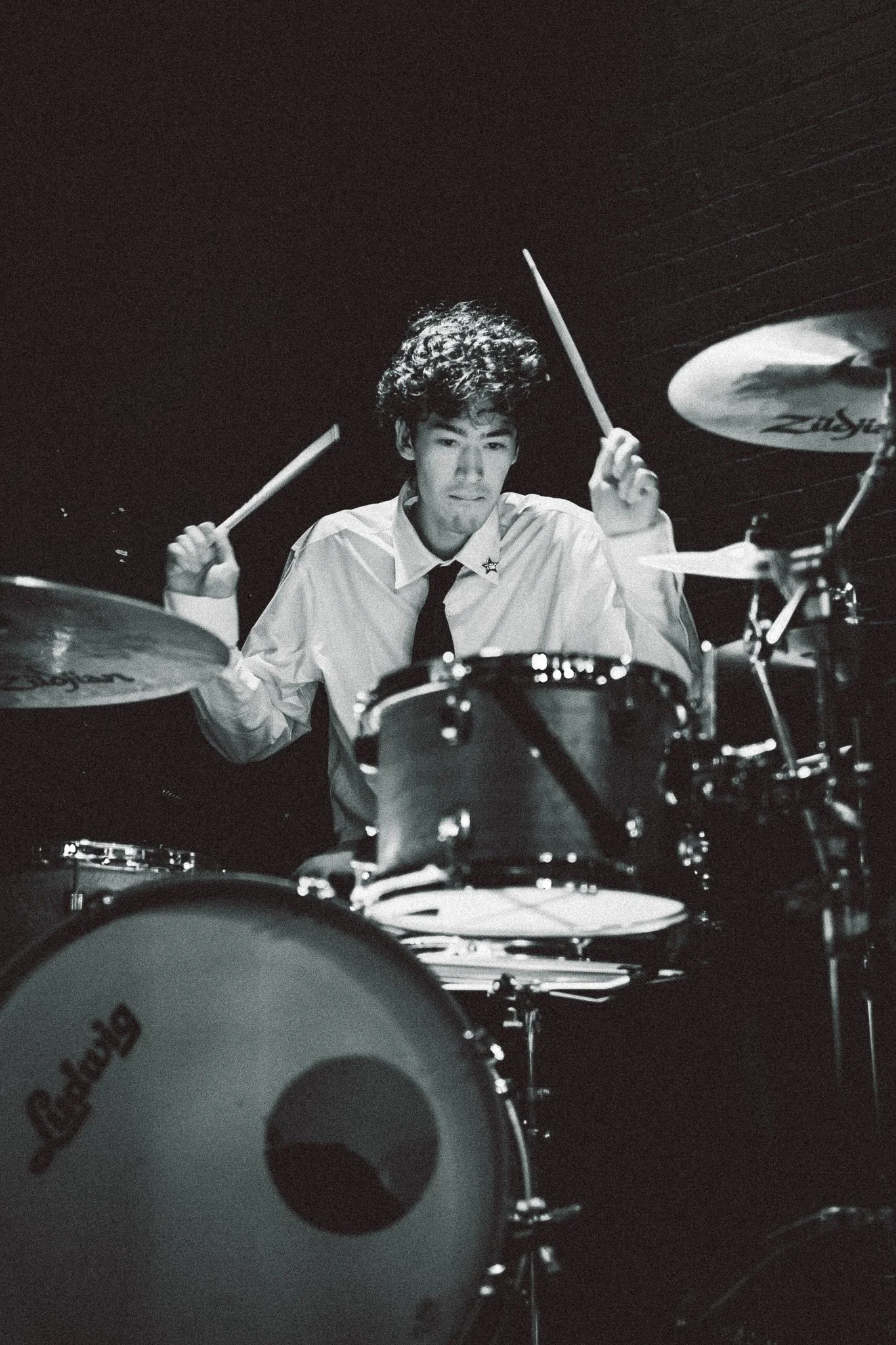 Black and white photo of a young man with curly hair playing a drum set, wearing a collared shirt and tie, against a dark background.