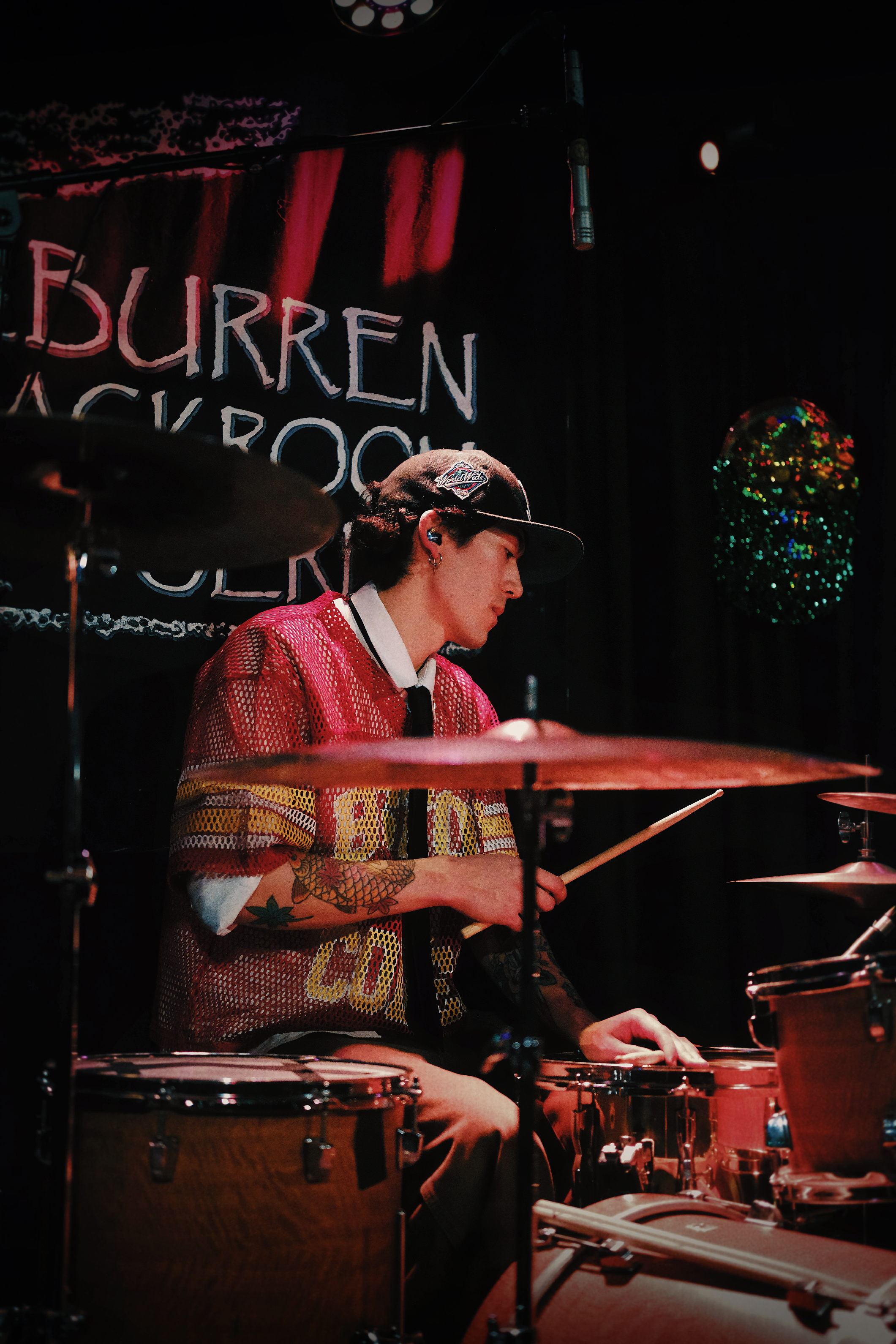 A person wearing a black cap and red mesh shirt playing drums on stage, with a black background and a colorful illuminated sign that reads 'BURR...