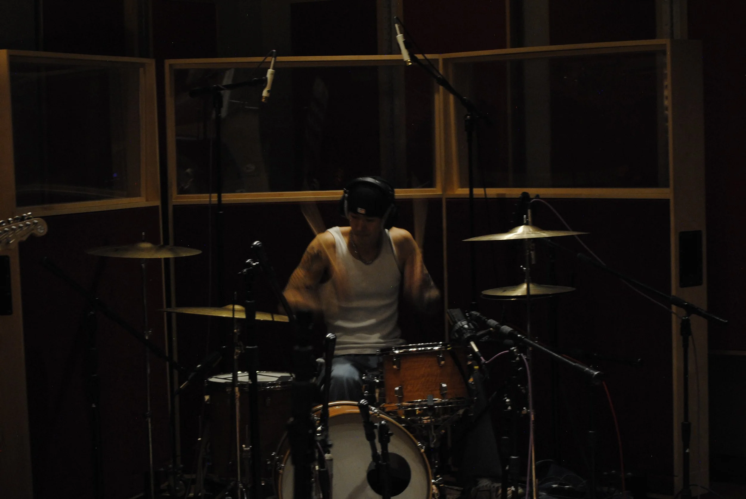 A person wearing a white tank top, headphones, and a baseball cap is sitting at a drum set in a recording studio. Microphones are positioned around them, and soundproof glass panels are in the background.