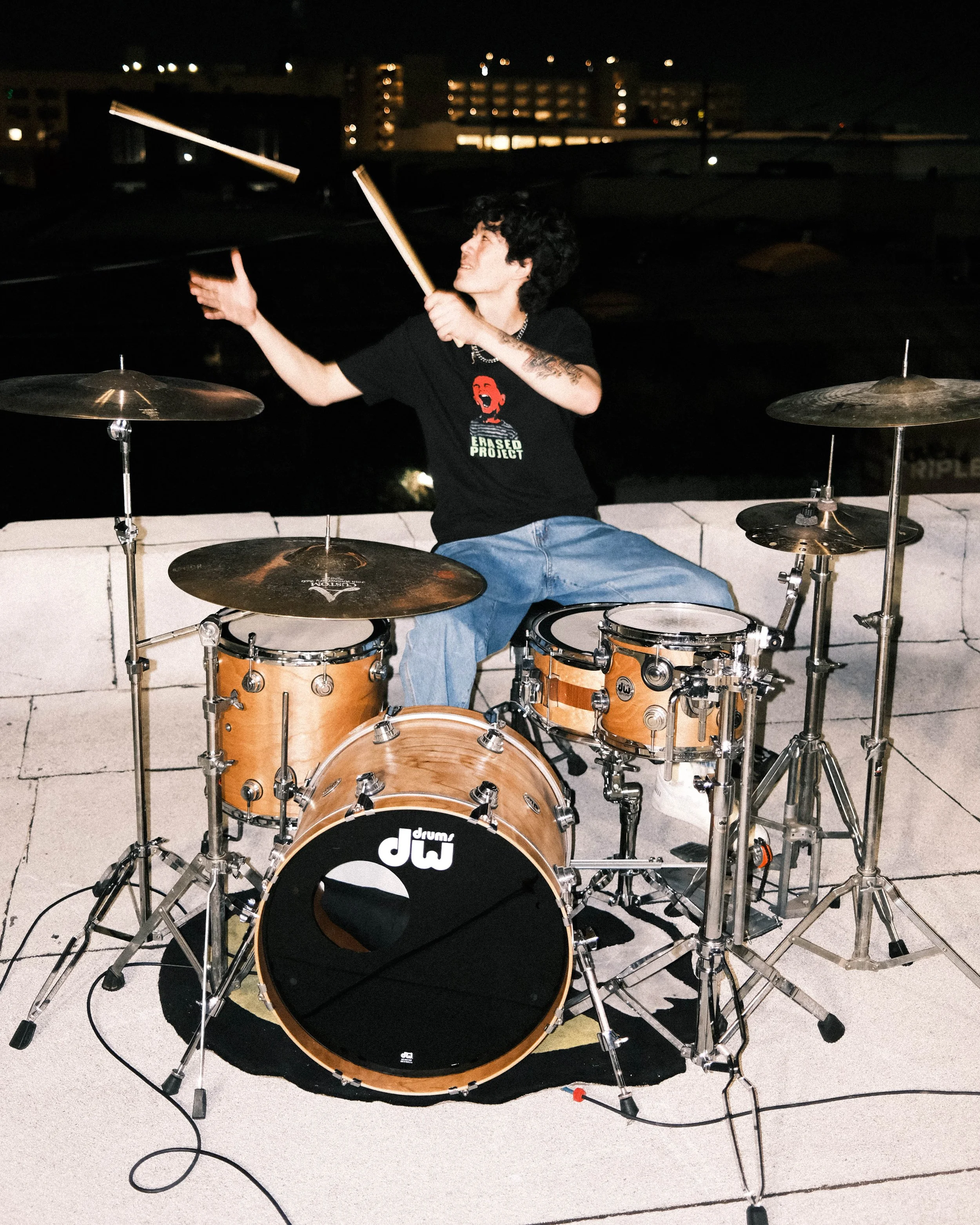 A young person with curly dark hair playing a drum set on a balcony at night, wearing a black T-shirt with a graphic design and a silver necklace. The drum set includes a bass drum, snare, toms, and cymbals, and is positioned on a white tiled surface with a black circular rug underneath.