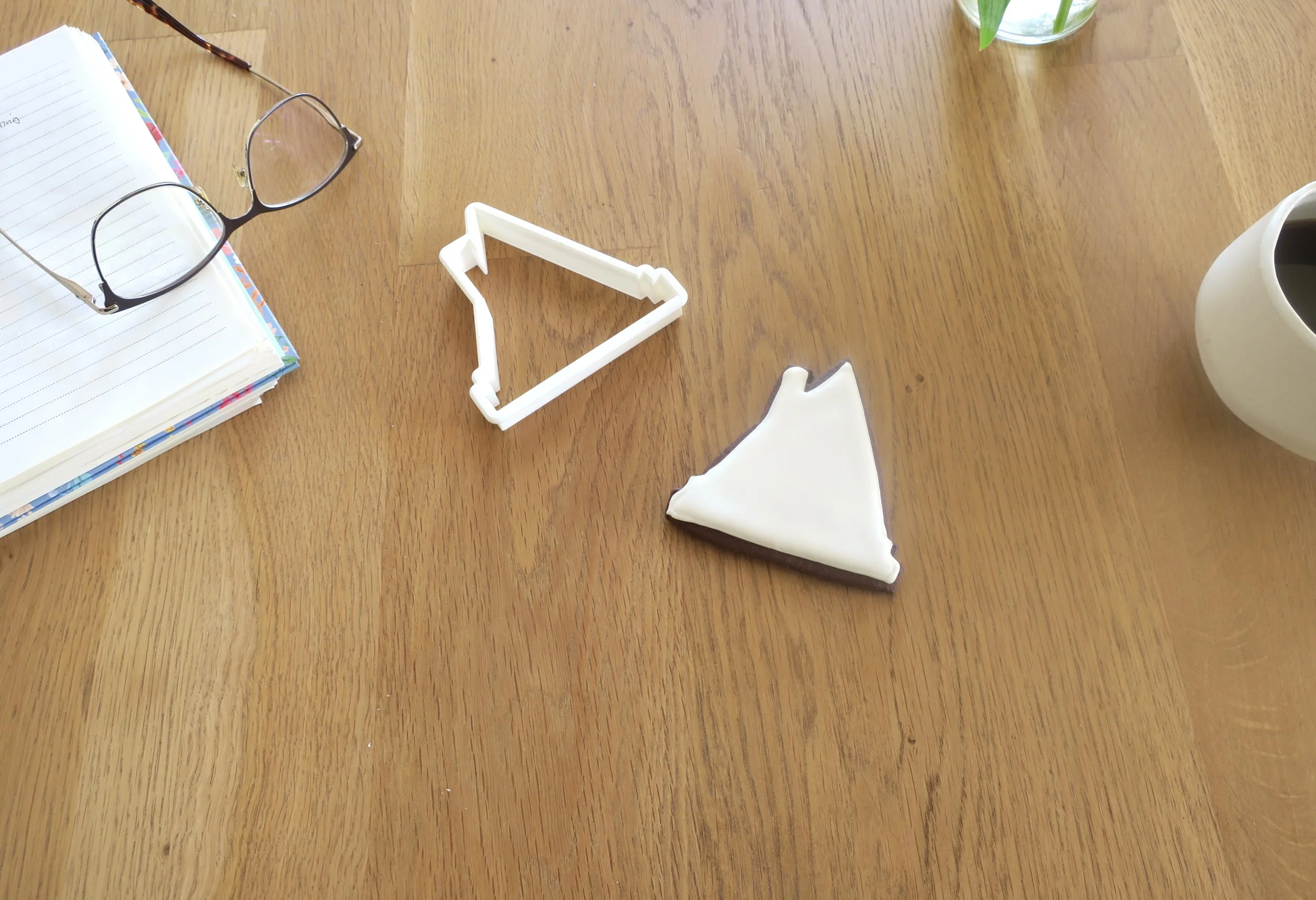 A wooden table with a stack of notebooks, a pair of glasses, a white plastic triangle slicer, a sheet of paper, a glass of water with green leaves, and a white mug of coffee.