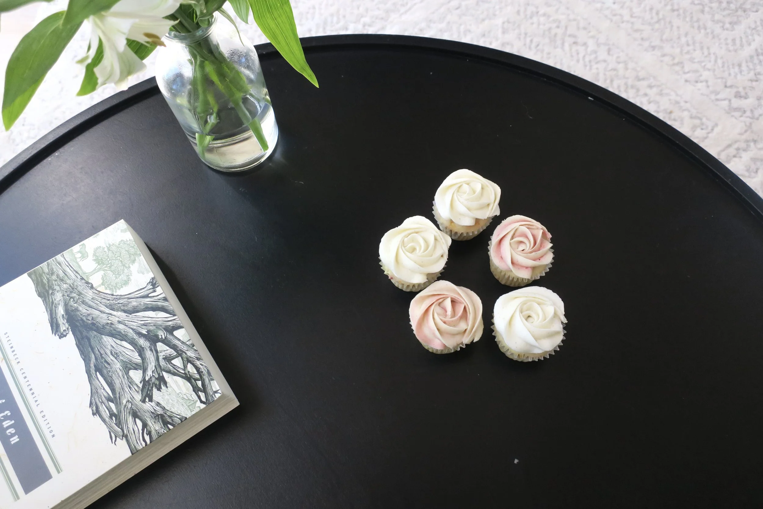 A black round table with a vase of green leaves and white flowers, and several cupcakes decorated to look like roses in pink and white. A book with a black and white tree illustration is partially visible in the lower left corner.