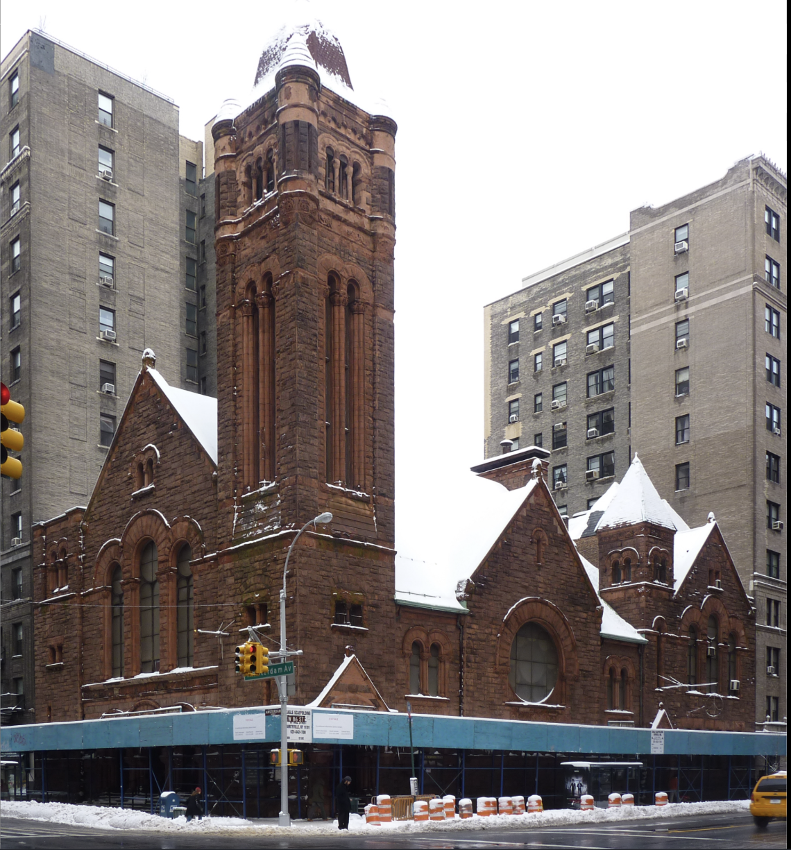 A historic red brick church with tall steeples and arched windows, snow-covered roof, surrounded by modern high-rise buildings in an urban setting.