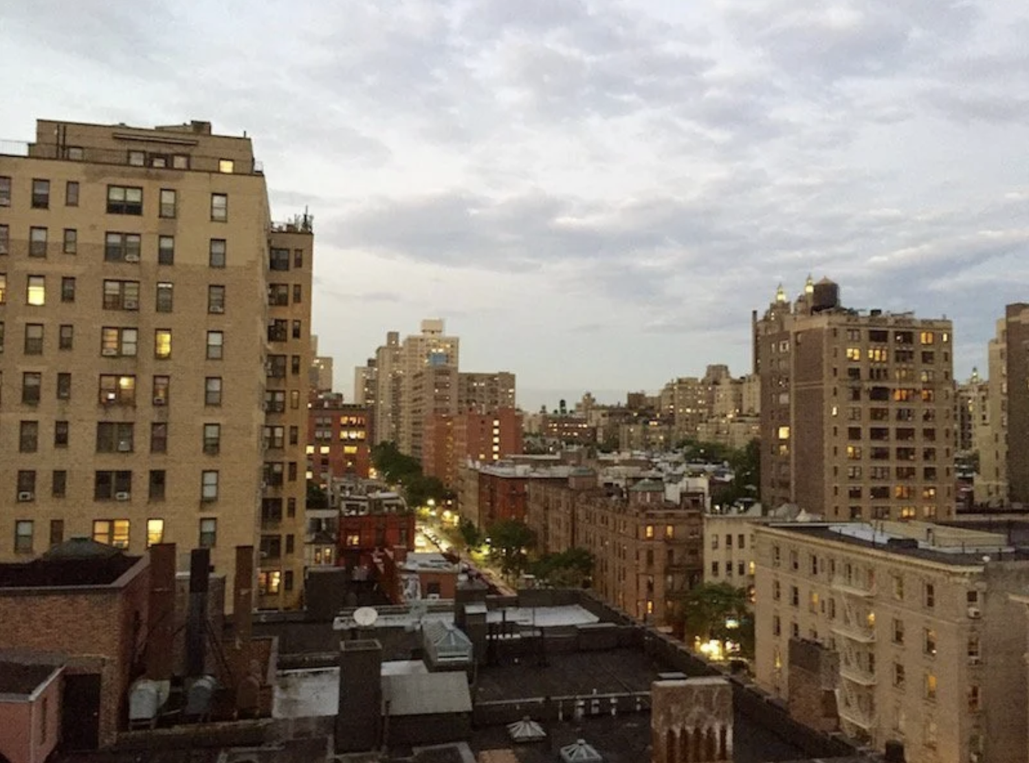 Cityscape with tall residential buildings and smaller structures, evening sky with clouds, some windows illuminated, streets with lights and trees.