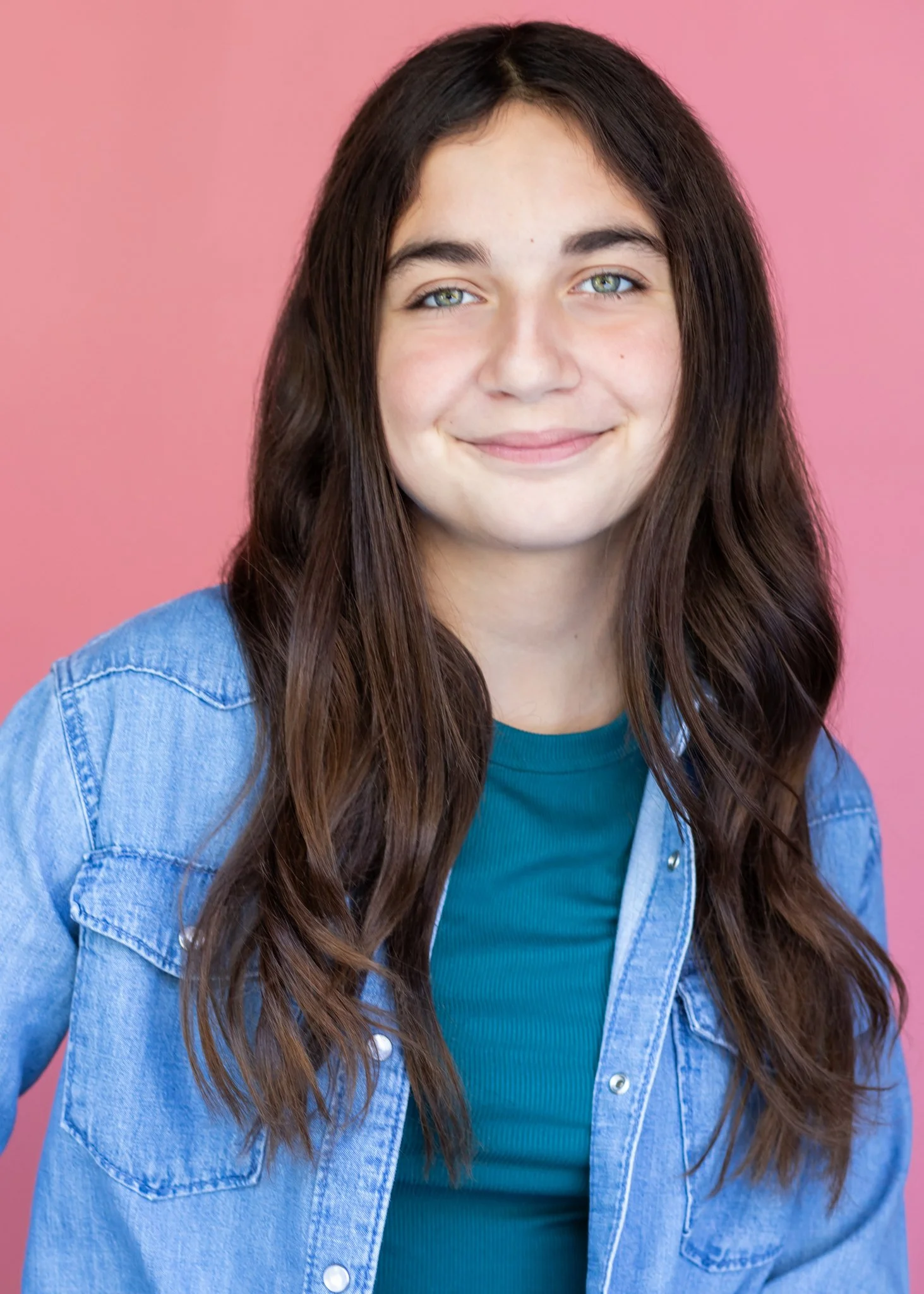 Close-up of a young woman with long brown hair, blue eyes, wearing a denim jacket and a teal top, smiling against a pink background.
