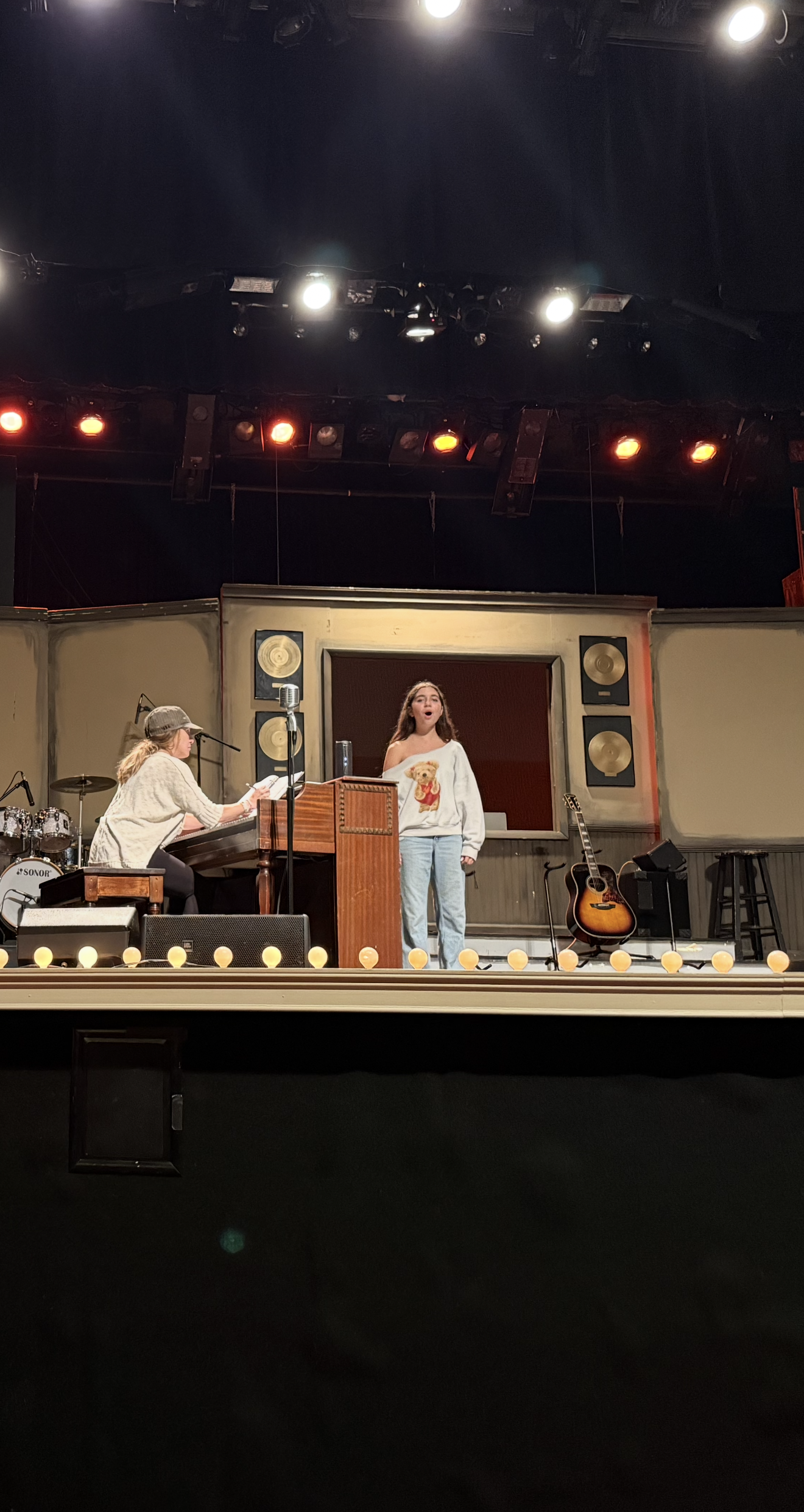 A woman singing on stage with a pianist playing a keyboard, a guitar on a stand, and stage lights overhead.