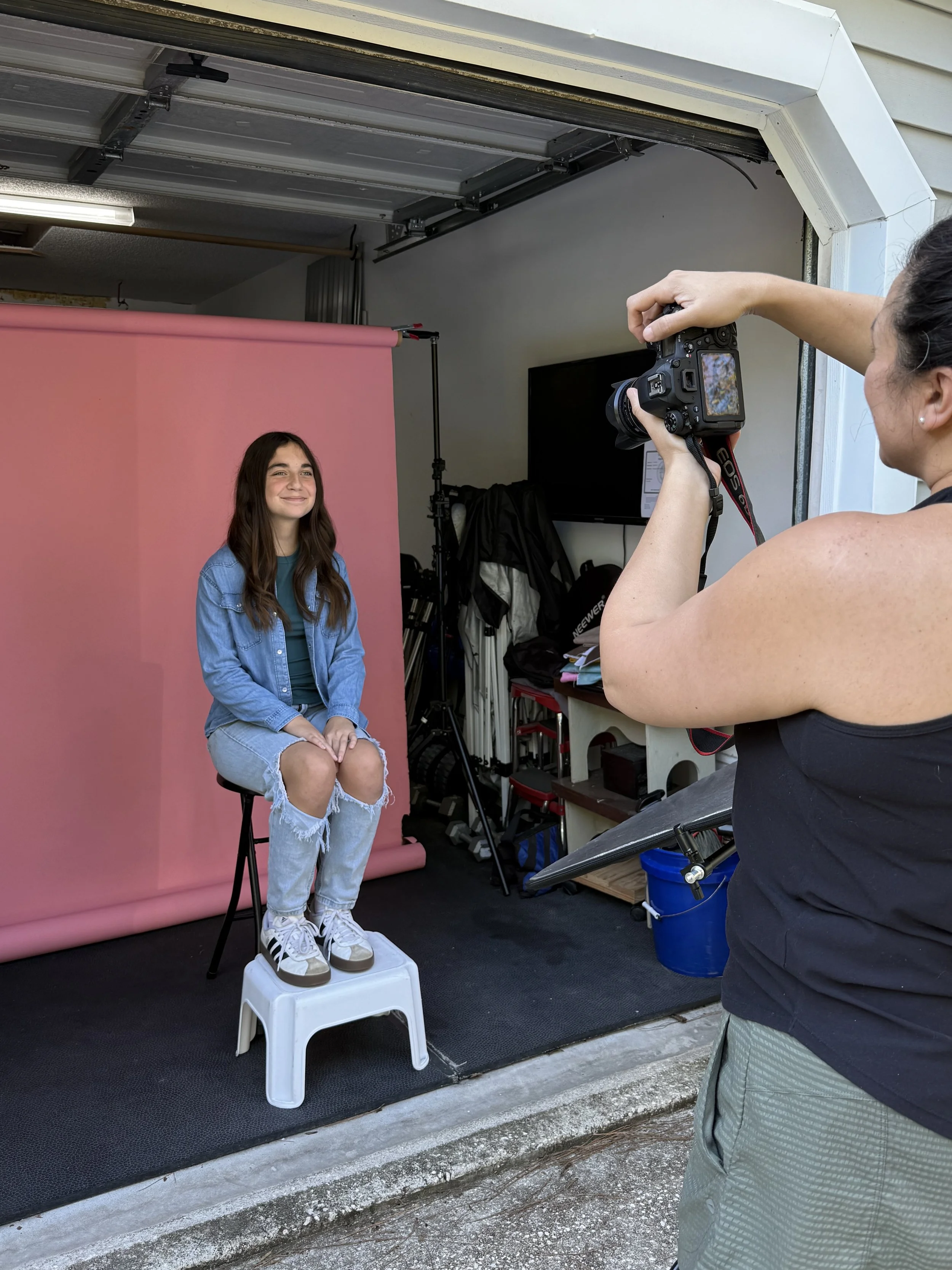 A woman sitting on a stool in front of a pink backdrop in a garage-like space, with a photographer taking her picture. The woman is smiling, wearing a denim jacket and ripped jeans, with sneakers, and the photographer is holding a camera with a display screen visible.