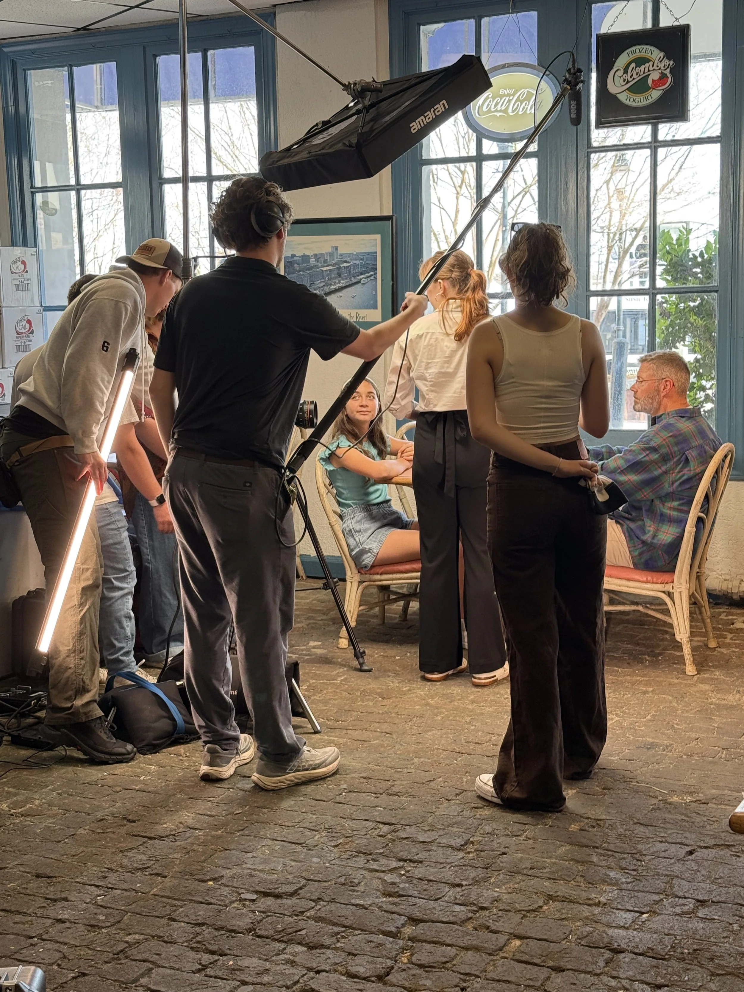 A behind-the-scenes photo of a film or photography shoot inside a cozy cafe or restaurant with large blue-framed windows. A group of people, including a woman sitting at a table and a man sitting across from her, are being filmed or photographed by a crew. The crew members are holding professional lighting and camera equipment, and a person is adjusting the lighting. The atmosphere appears to be casual, with some individuals engaging in conversation or waiting for their turn.