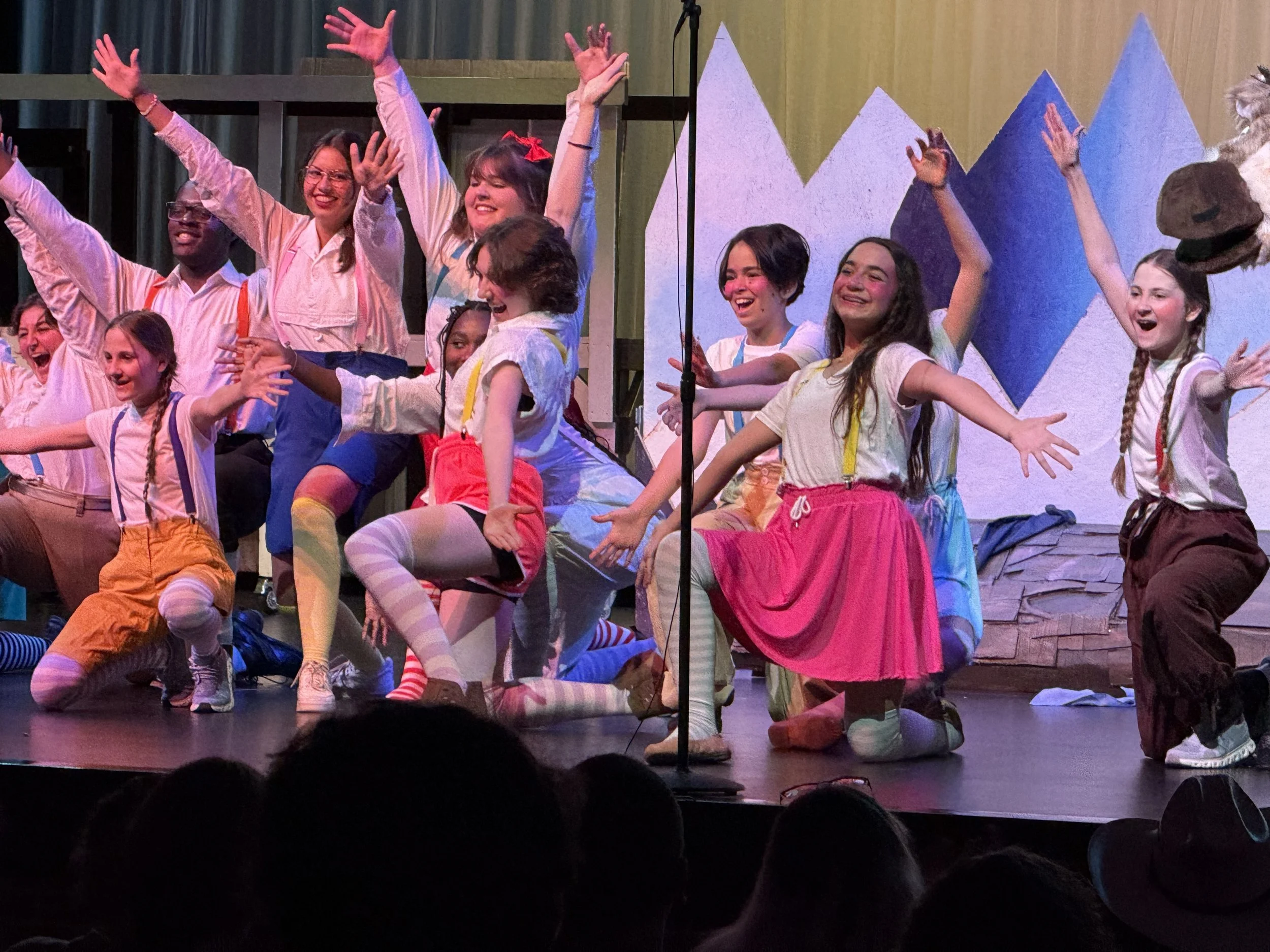 Group of children on stage performing a musical or play, wearing colorful costumes with painted stockings and suspenders, with a scenic mountain backdrop.