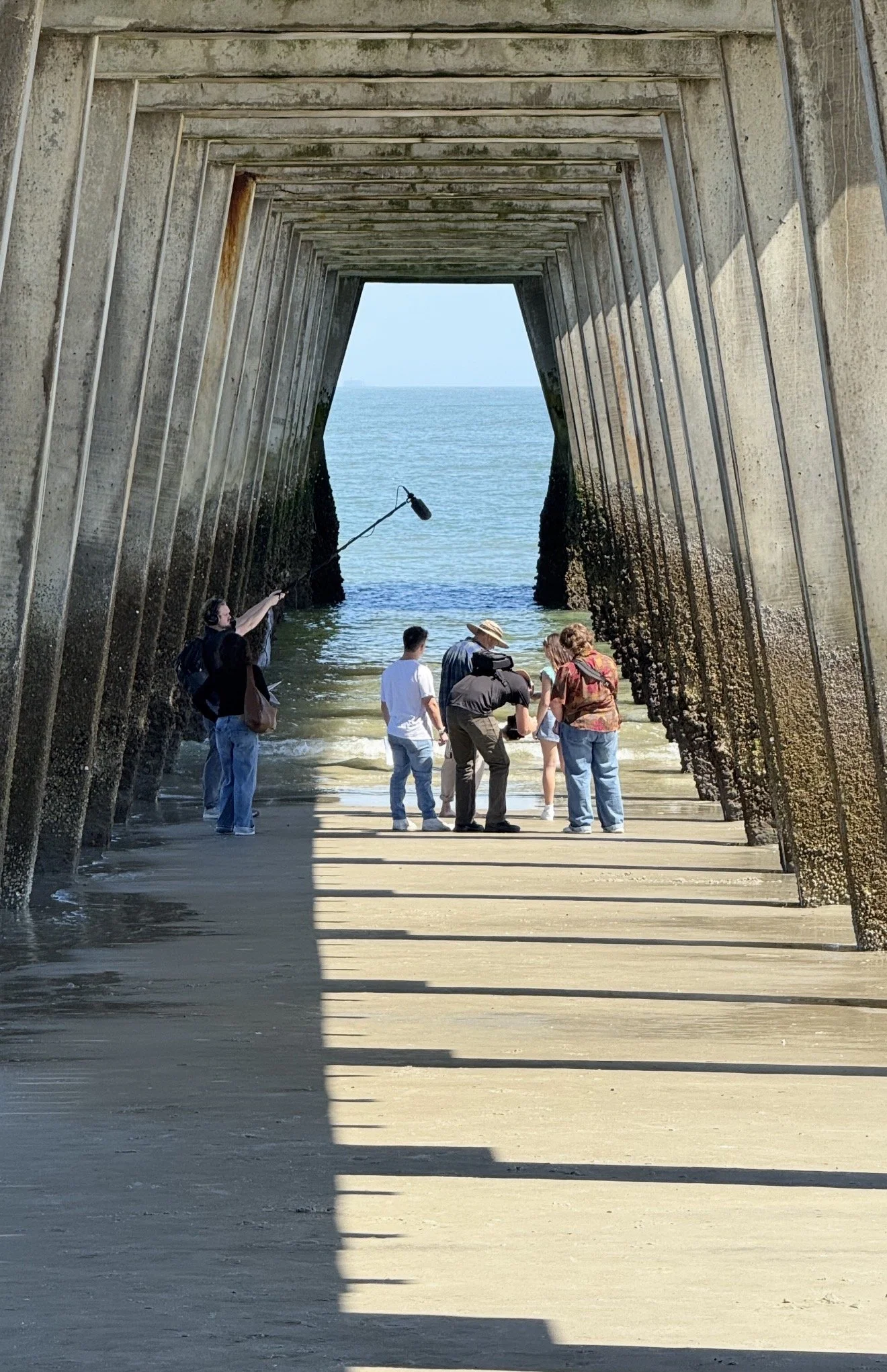 A group of people under a concrete pier by the water, with one person holding a boom microphone, capturing audio during a film or photo shoot.