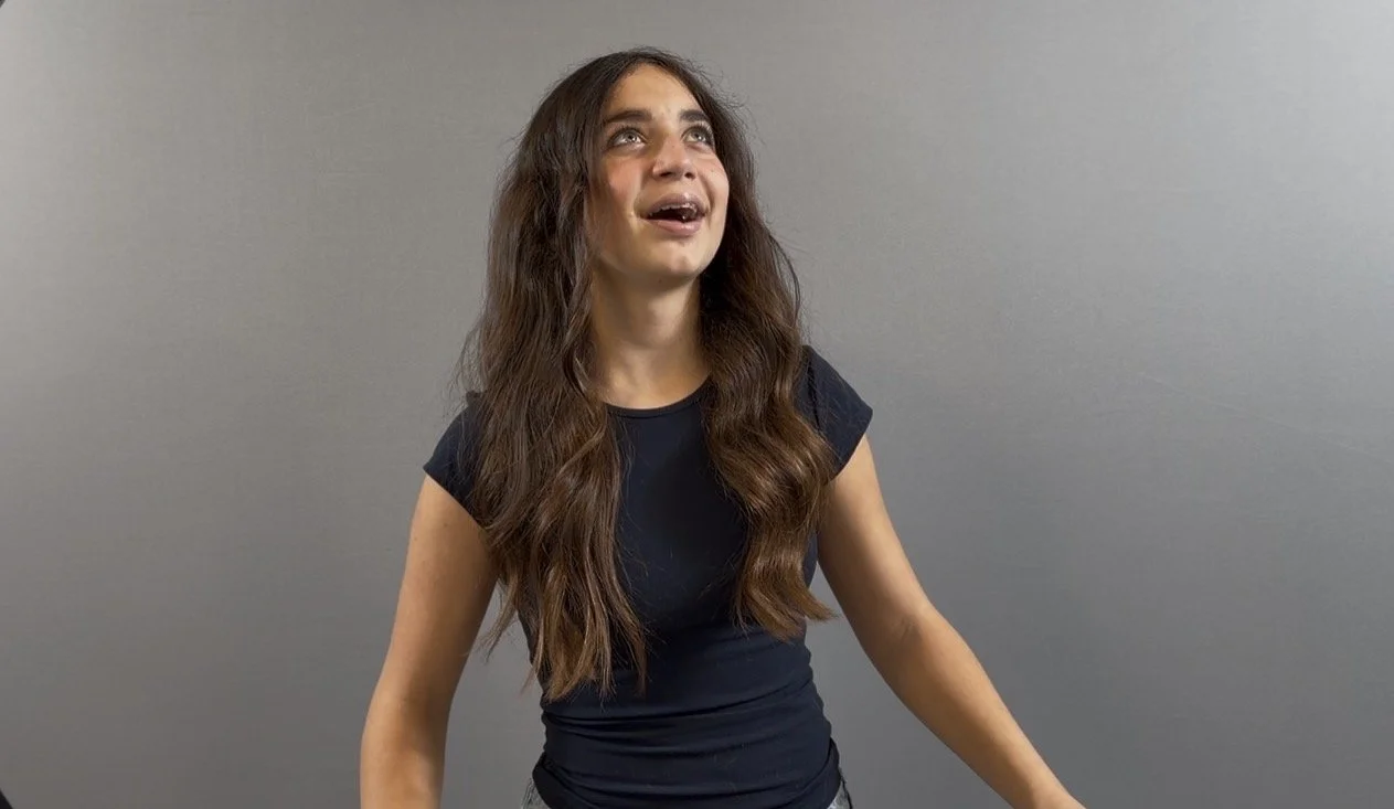 Young woman with long, wavy brown hair looking upward with a surprised or excited expression, wearing a black T-shirt, standing against a plain gray background.