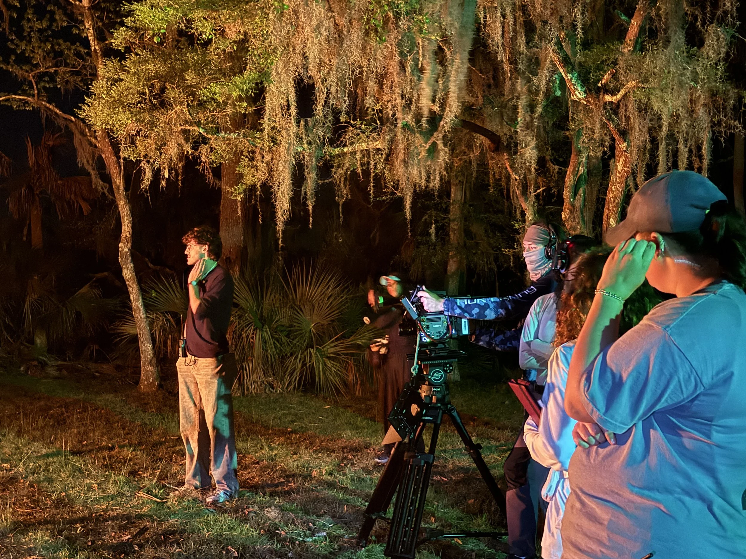 Night scene with people and a camera setup in a wooded area, illuminated by colorful lights.