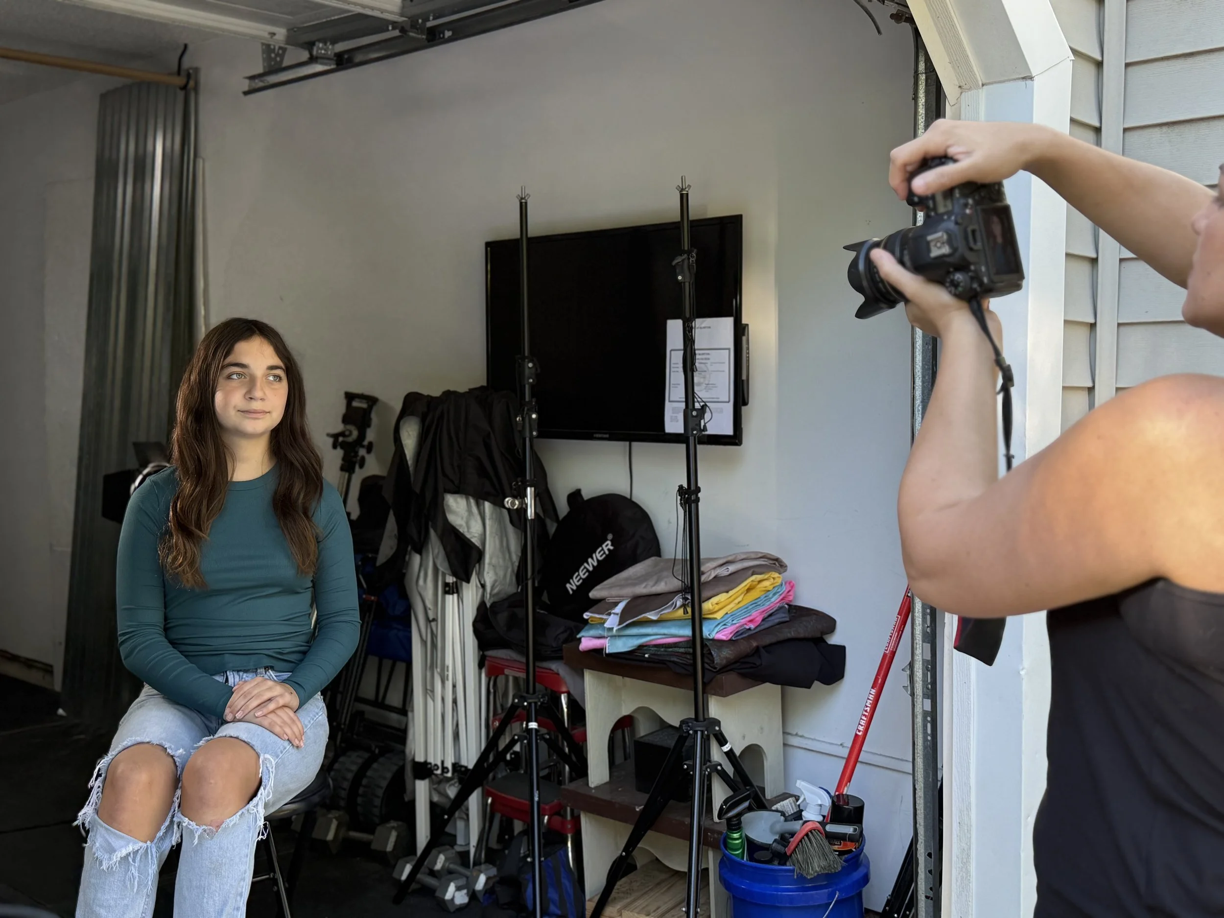 A woman with long brown hair, wearing a teal long sleeve shirt and ripped jeans, sitting on a black chair, having her photo taken by a photographer. The background includes a white wall, a flat-screen TV, and various photography equipment and gear.