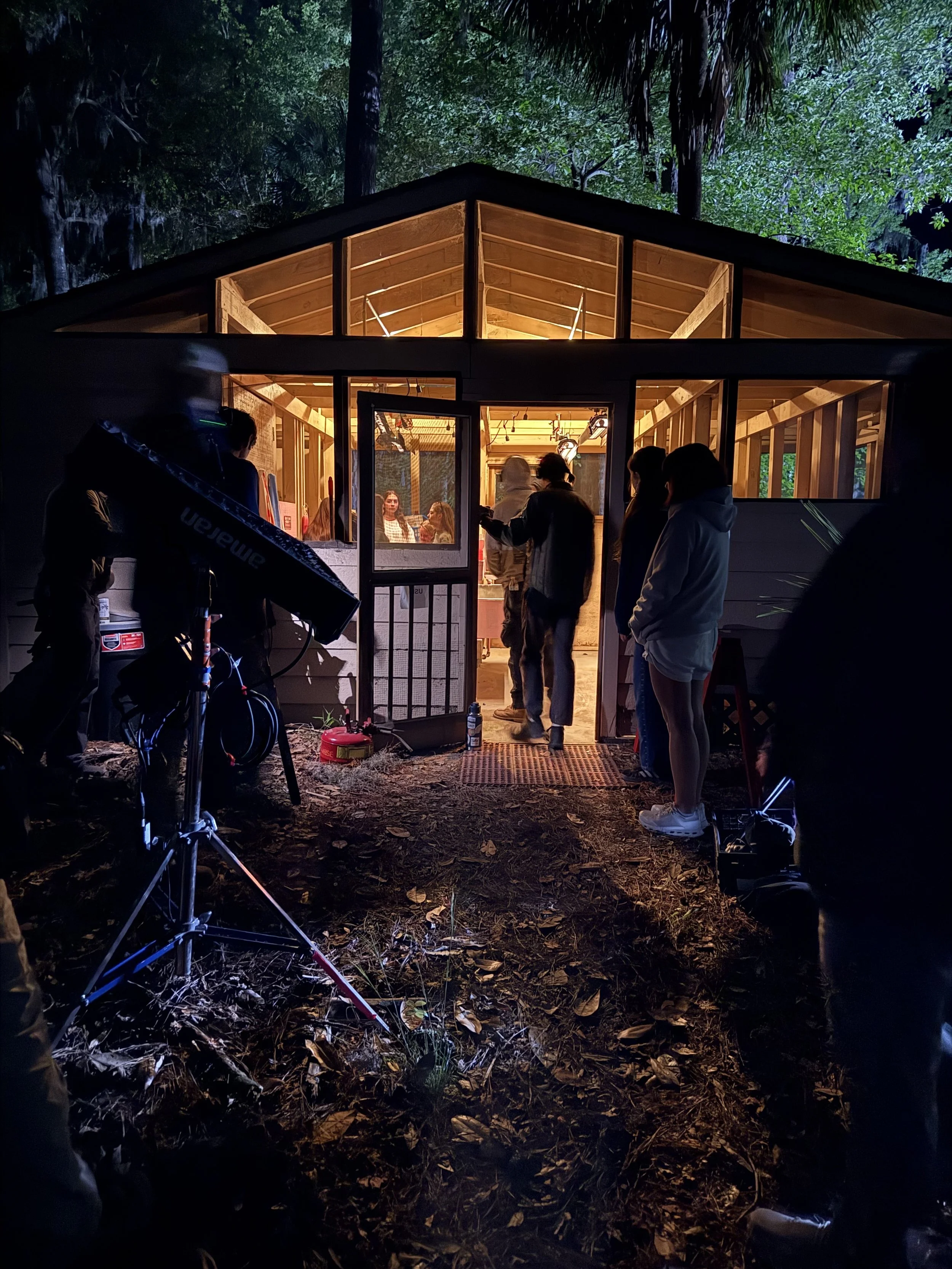 Scene of a film crew shooting inside a wooden cabin in a forest at night. Crew members are gathered outside, some looking in through the open door, with professional lighting equipment set up outside on the ground covered with leaves.