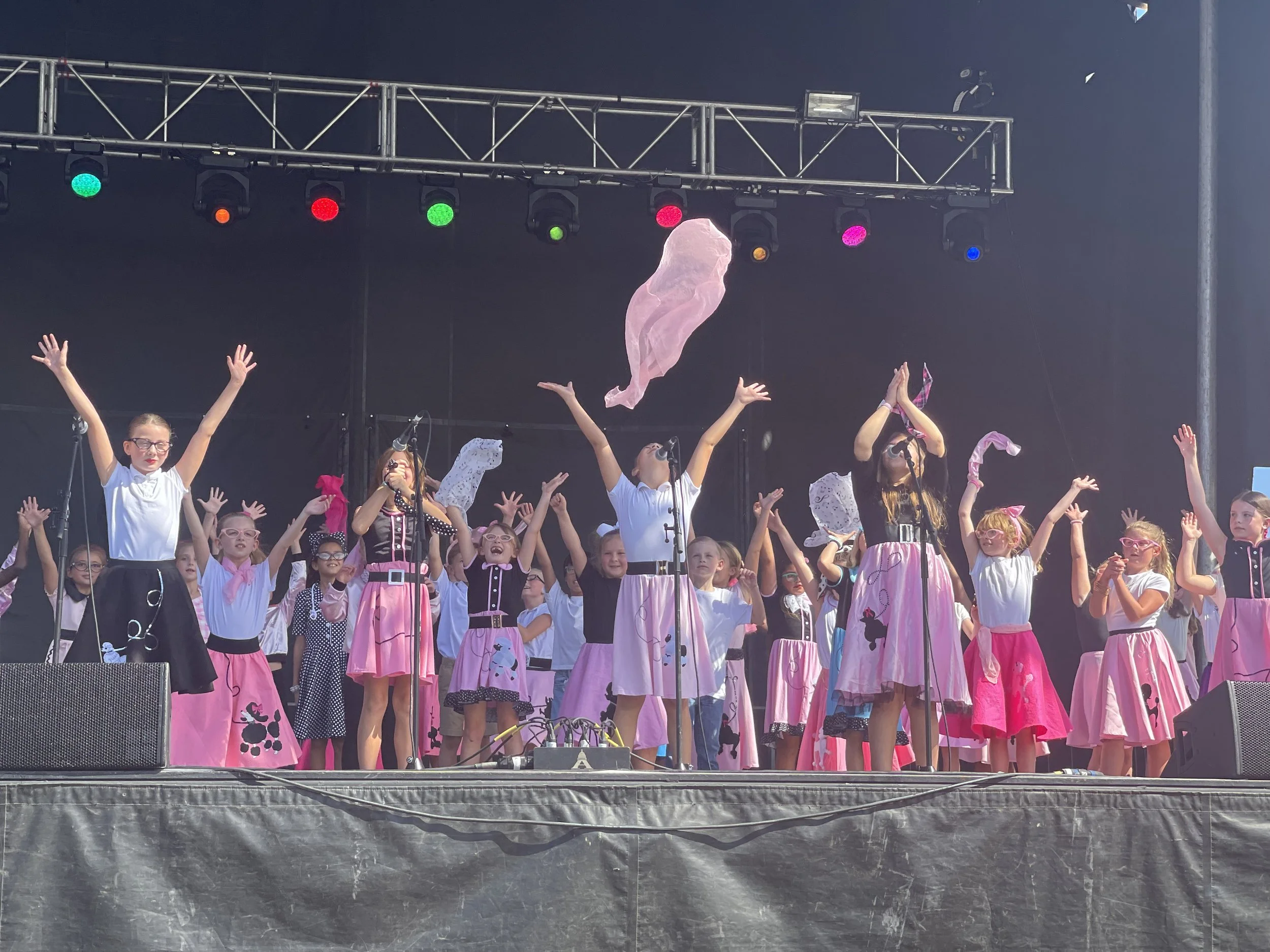 Children on stage performing with pink skirts with cow print and pink ribbons, raising their hands, with stage lights overhead.