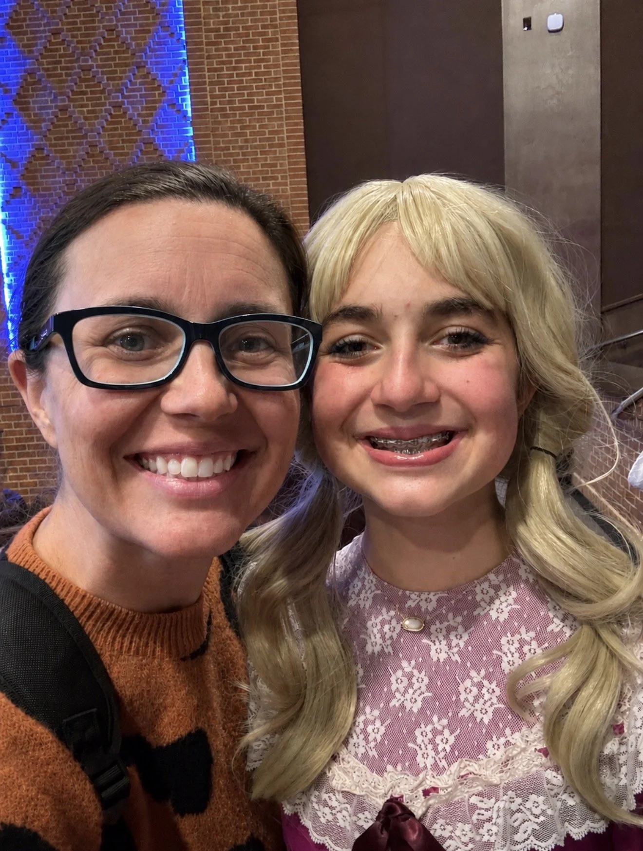 A woman with short dark hair, glasses, and a warm smile, and a young girl with long blonde hair, braces, and a lace dress, smiling for a selfie inside a building with brick and wooden walls.