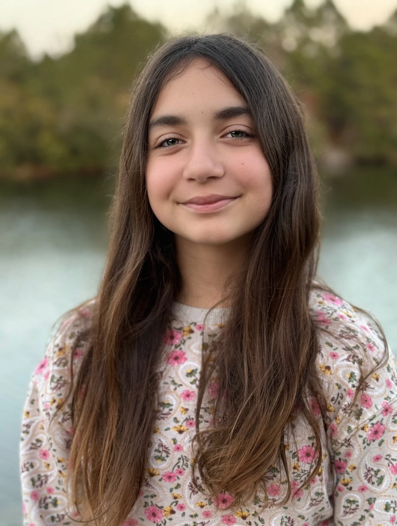 A young girl with long brown hair and gray eyes standing outdoors near a body of water with trees in the background, wearing a cream-colored floral shirt.