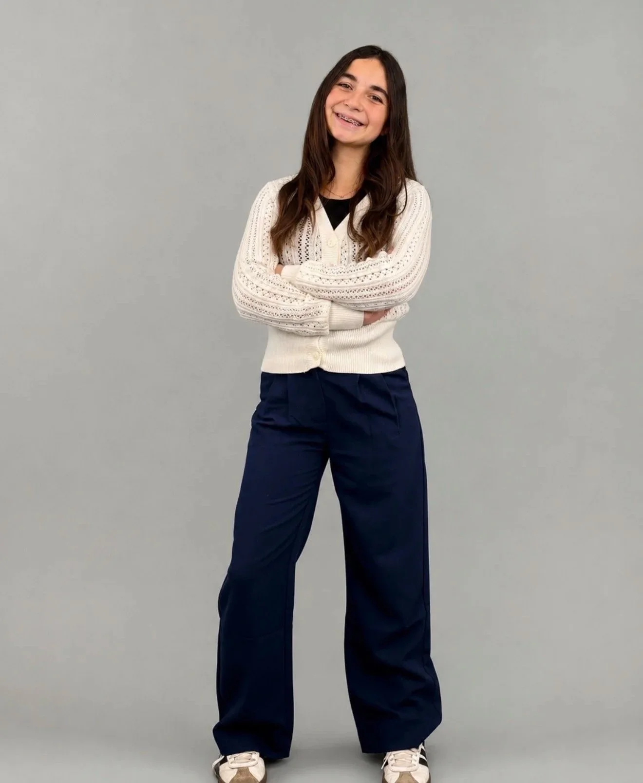 A young woman with long brown hair, wearing a white knitted sweater over a black shirt and navy blue wide-leg pants, standing with arms crossed and smiling against a plain light gray background.