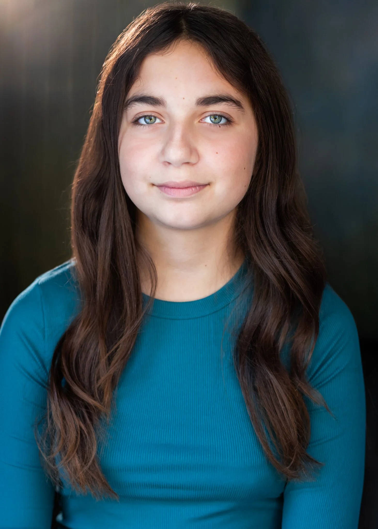 Close-up portrait of a young woman with long wavy brown hair and green eyes, smiling slightly, wearing a teal top, against a dark background.