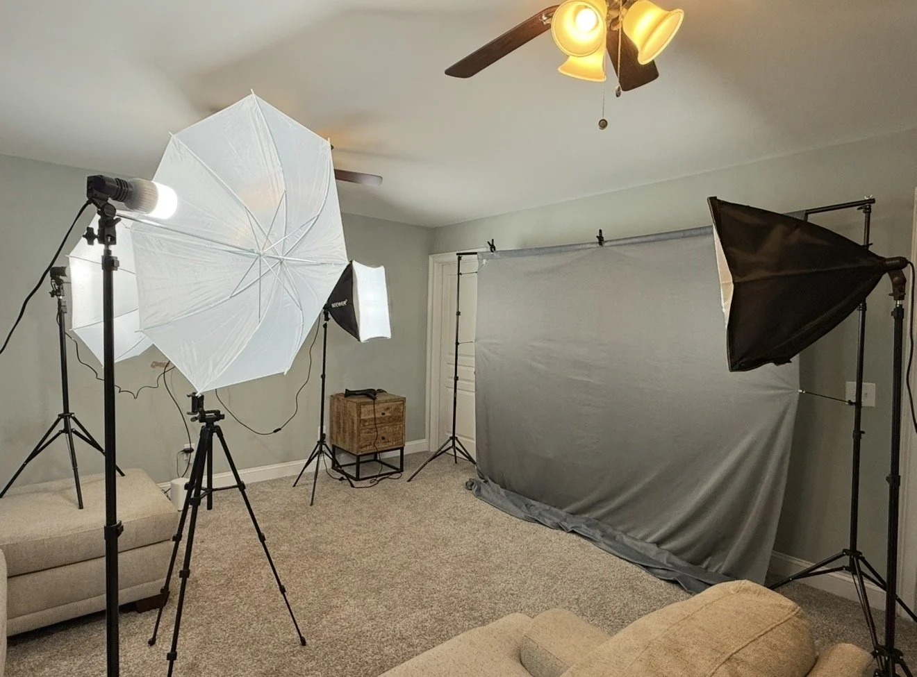 Home photography studio setup with four softbox lights, a large white umbrella light, a gray backdrop on a stand, a small wooden table, sofas, and a ceiling fan with lights in the background.