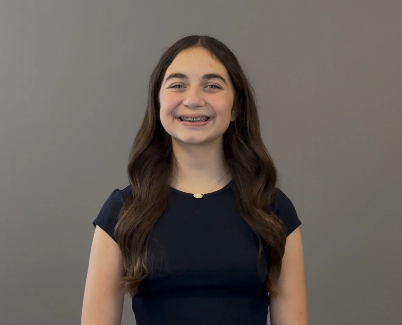 A young girl with long brown hair, smiling and showing braces, wearing a black shirt and a necklace, standing against a gray background.