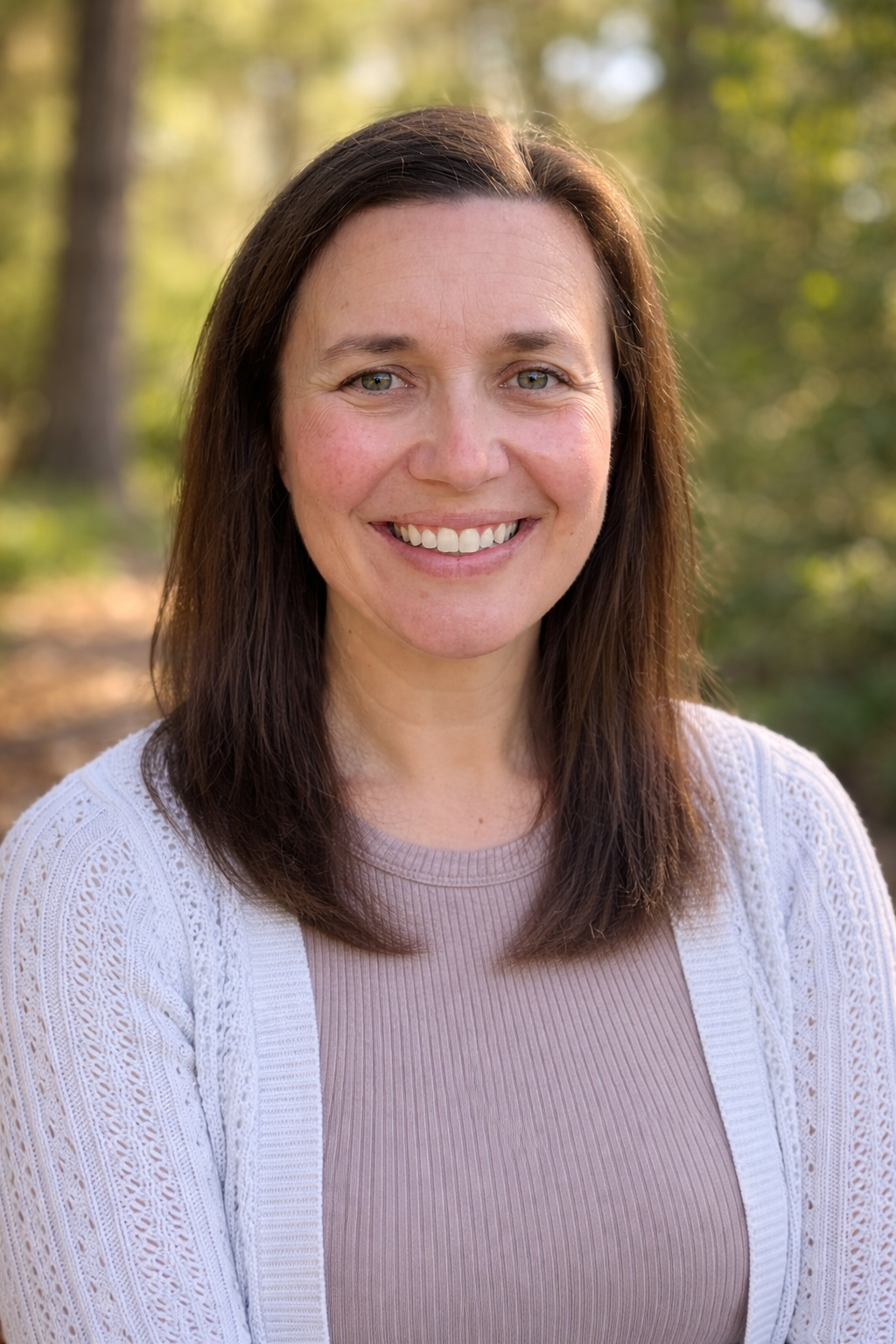 A smiling woman with brown hair and blue eyes standing outdoors in a forested area during daytime.