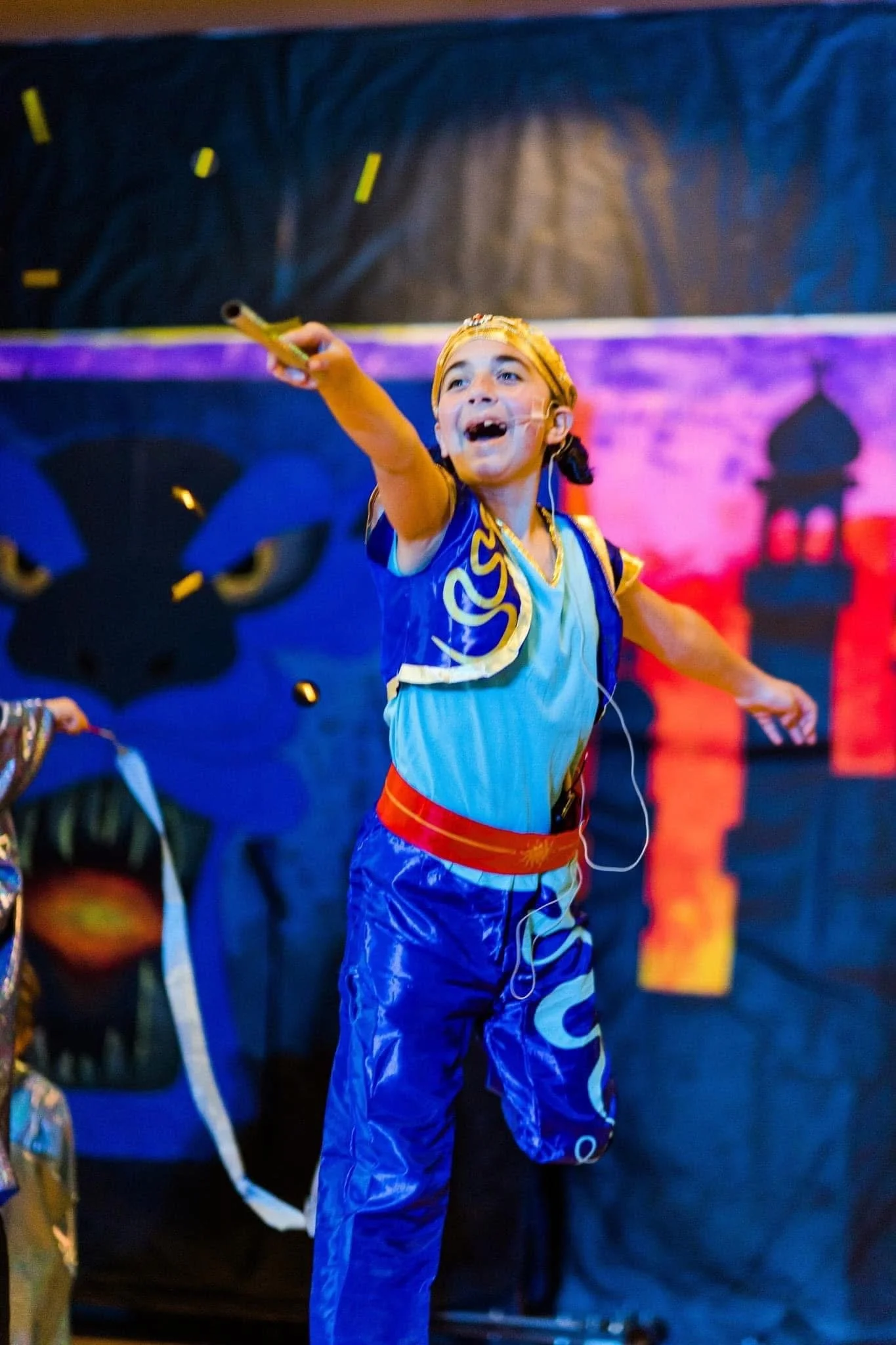 A young girl with a joyful expression, wearing traditional colorful clothing and a headband, pointing upwards while holding a flute. She is in a vibrant environment with a backdrop featuring silhouettes of structures and colors that suggest an outdoor or cultural celebration.