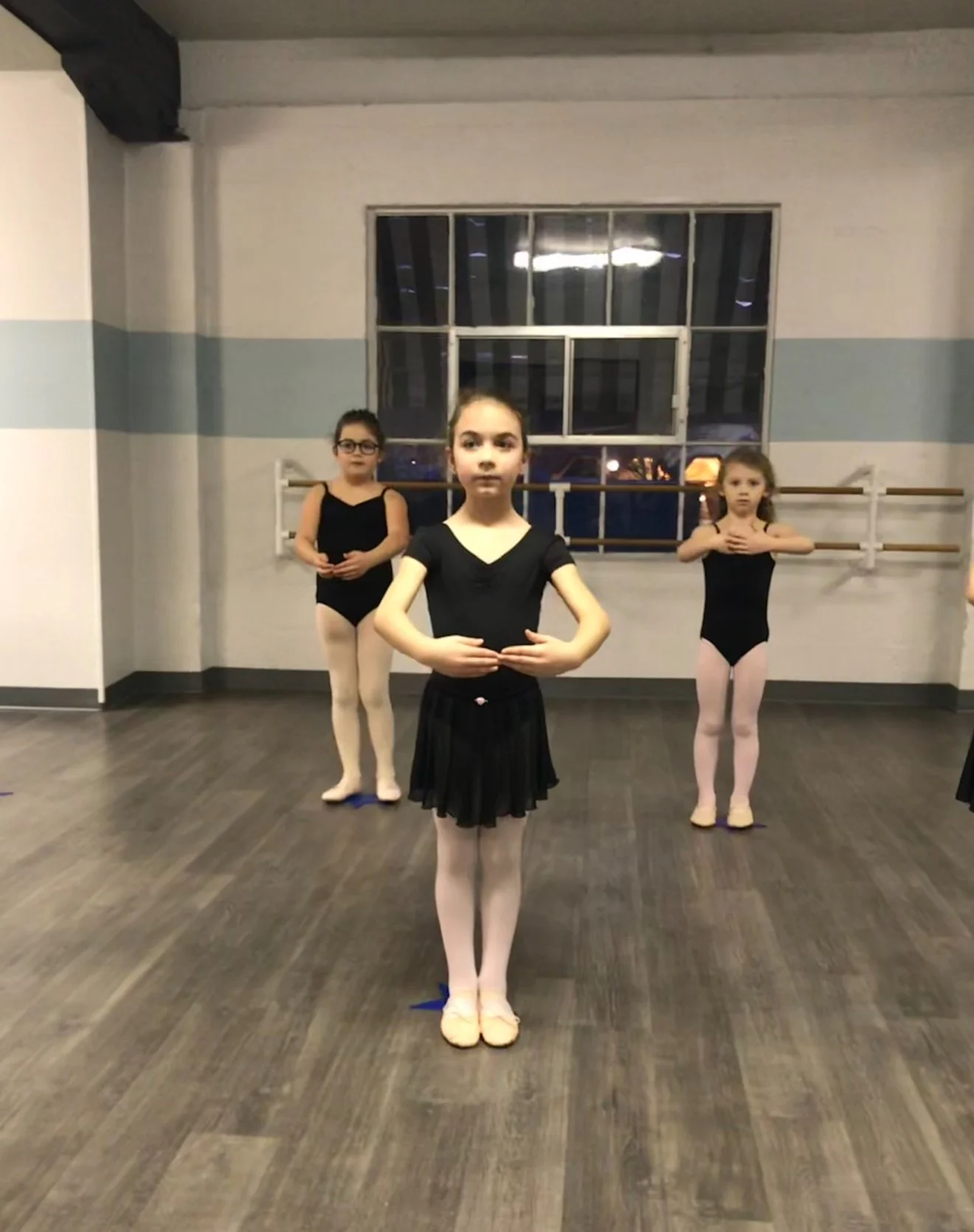 Three young girls dressed in ballet attire standing in ballet studio with wooden floor, mirror, ballet barres, and a large window at the back, posing for ballet practice.