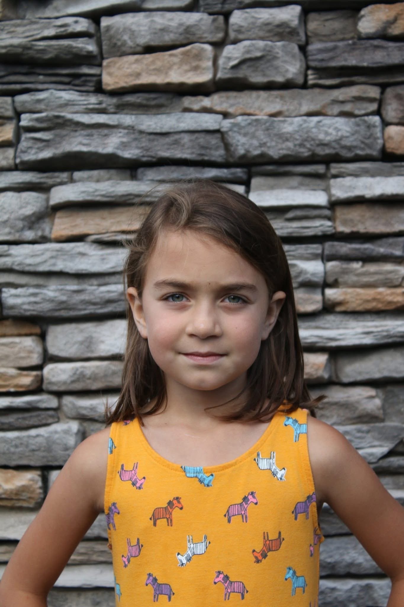A young girl with brown hair and blue eyes standing in front of a stone wall, wearing a yellow sleeveless top with colorful zebra patterns.