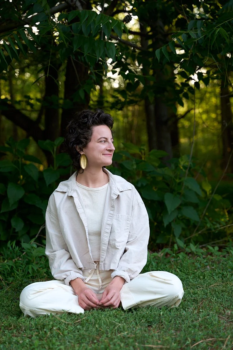 A woman sitting cross-legged on grass outdoors, surrounded by green foliage, smiling as she looks to her left.