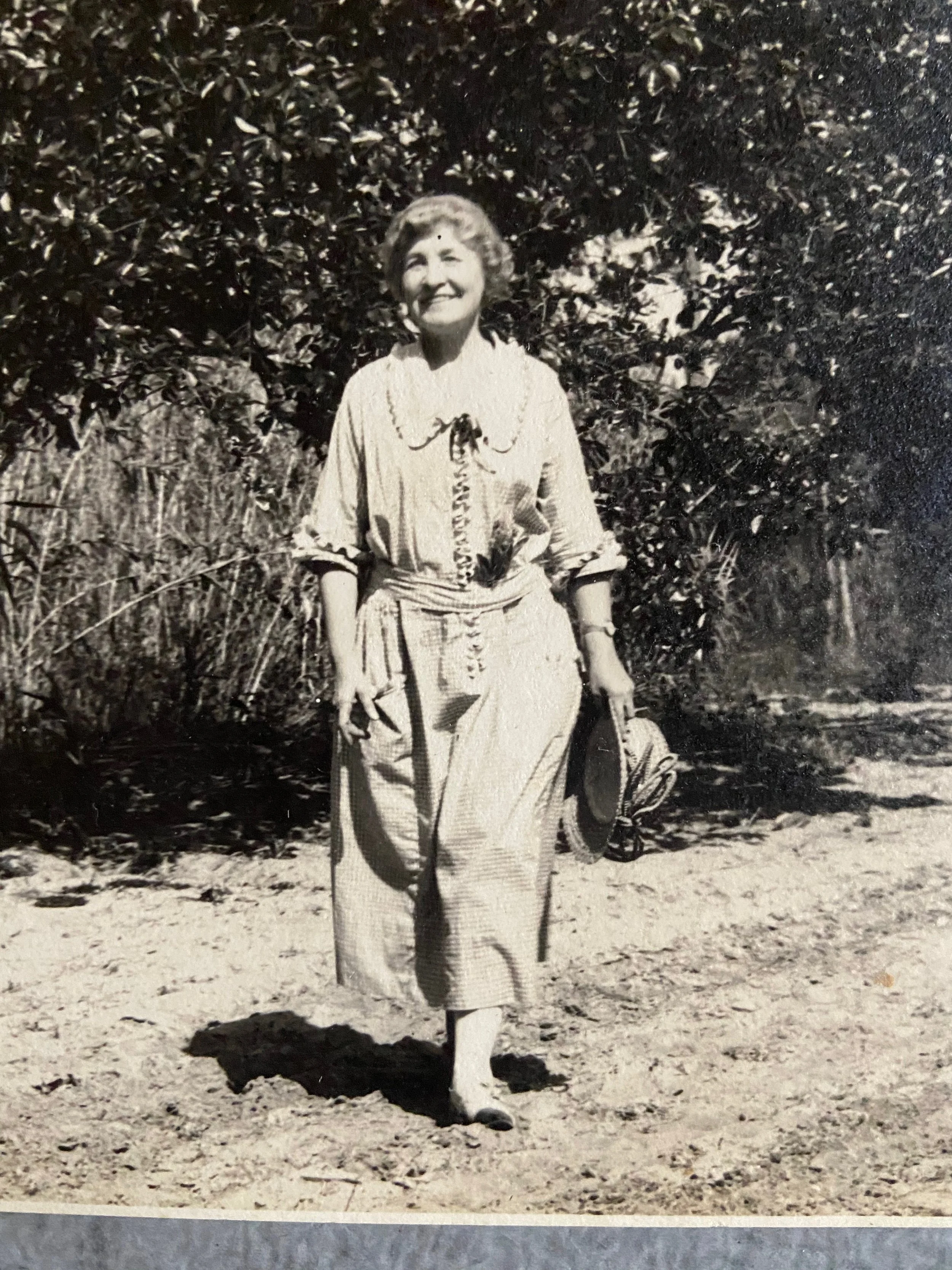 A woman stands in a sandy outdoor area, smiling, dressed in a patterned dress with a necklace, holding a walking stick, with a large tree or bush behind her.