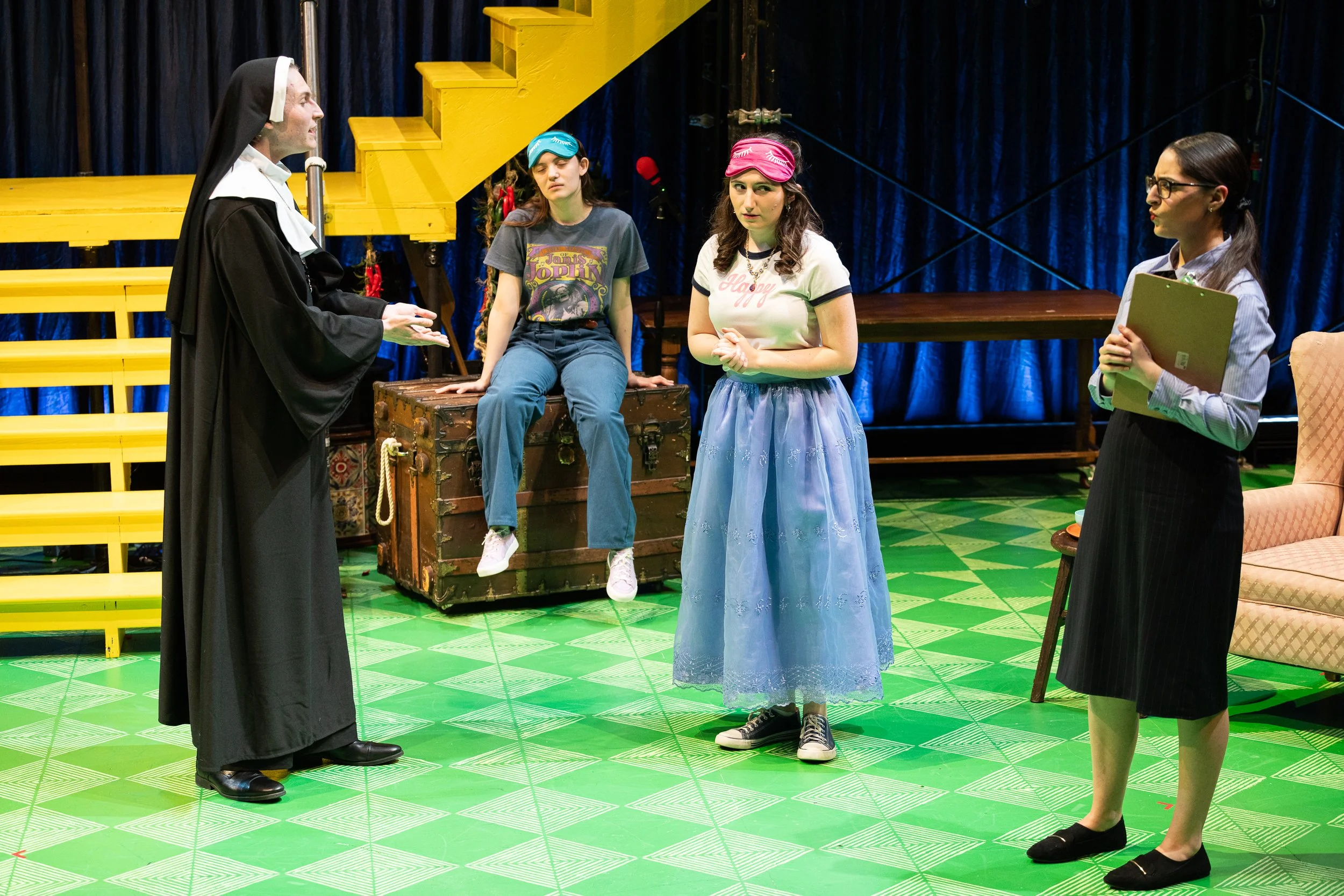A theatrical scene with four women on stage. One dressed as a nun speaking to three more women, one sitting on a trunk wearing a t-shirt and jeans, another wearing a pink headband and a white t-shirt, and the third in a suit holding a clipboard. The 