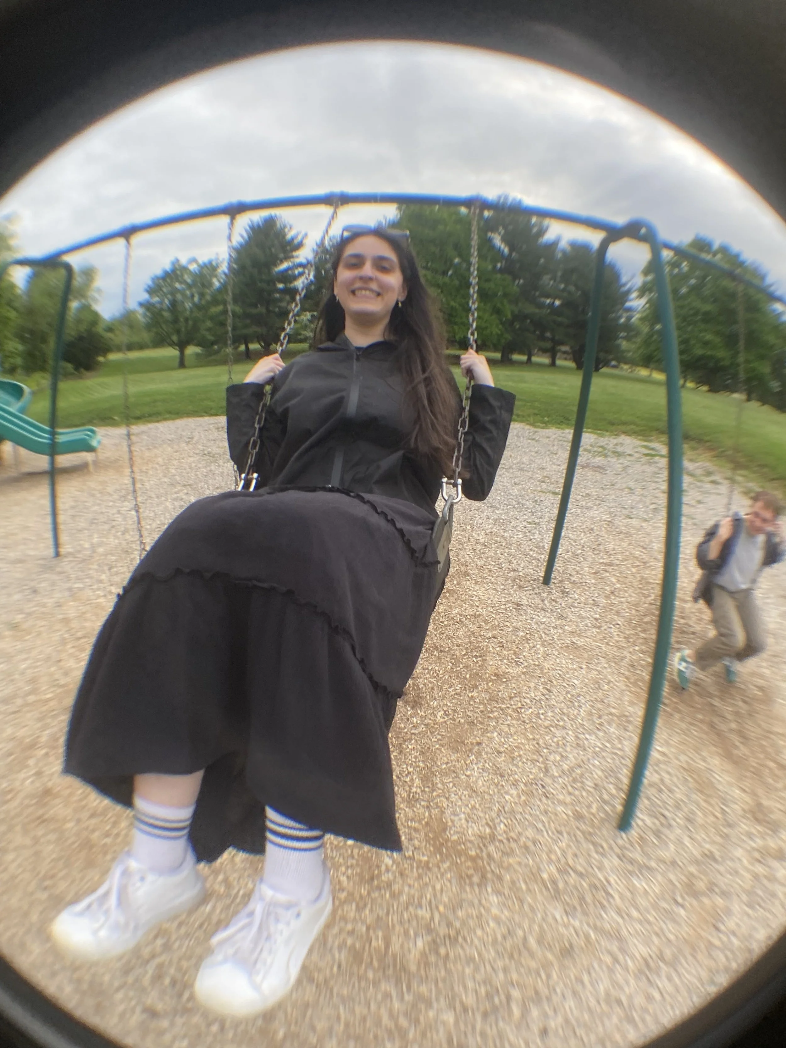 A girl smiling and sitting on a swing in a park, wearing a black jacket, black skirt, white sneakers, and striped socks. There are trees and a slide in the background, with a cloudy sky overhead.