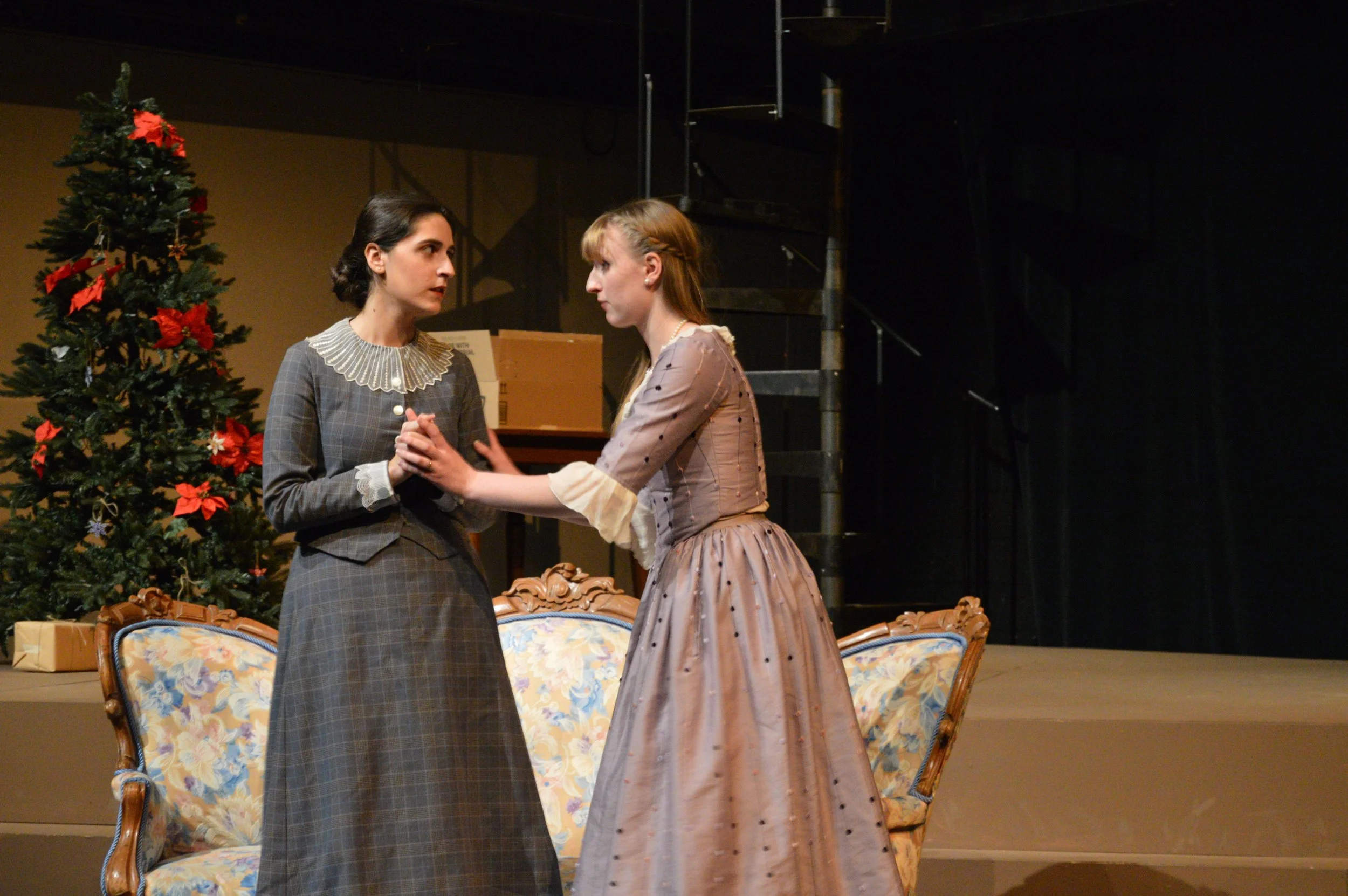 Two women dressed in vintage clothing are standing close to each other, facing and holding hands, in front of a decorated Christmas tree on a stage. There are boxes and chairs behind them, and a staircase to the right.