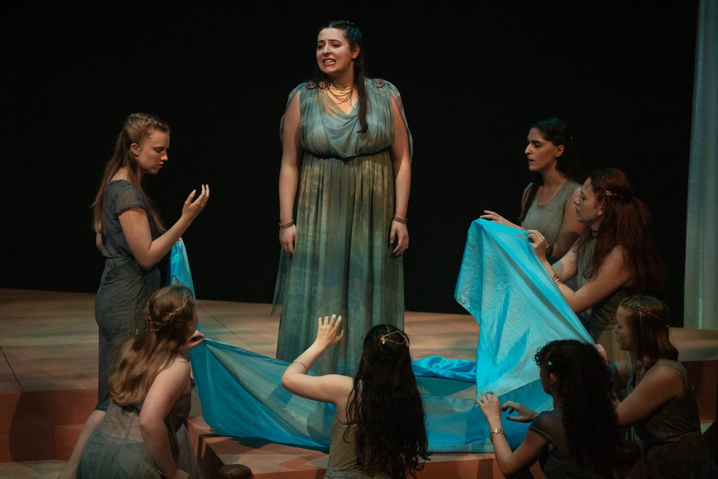 Women on stage, with one standing in the center, wearing a flowing dress, surrounded by others holding blue fabric, performing a scene or act.