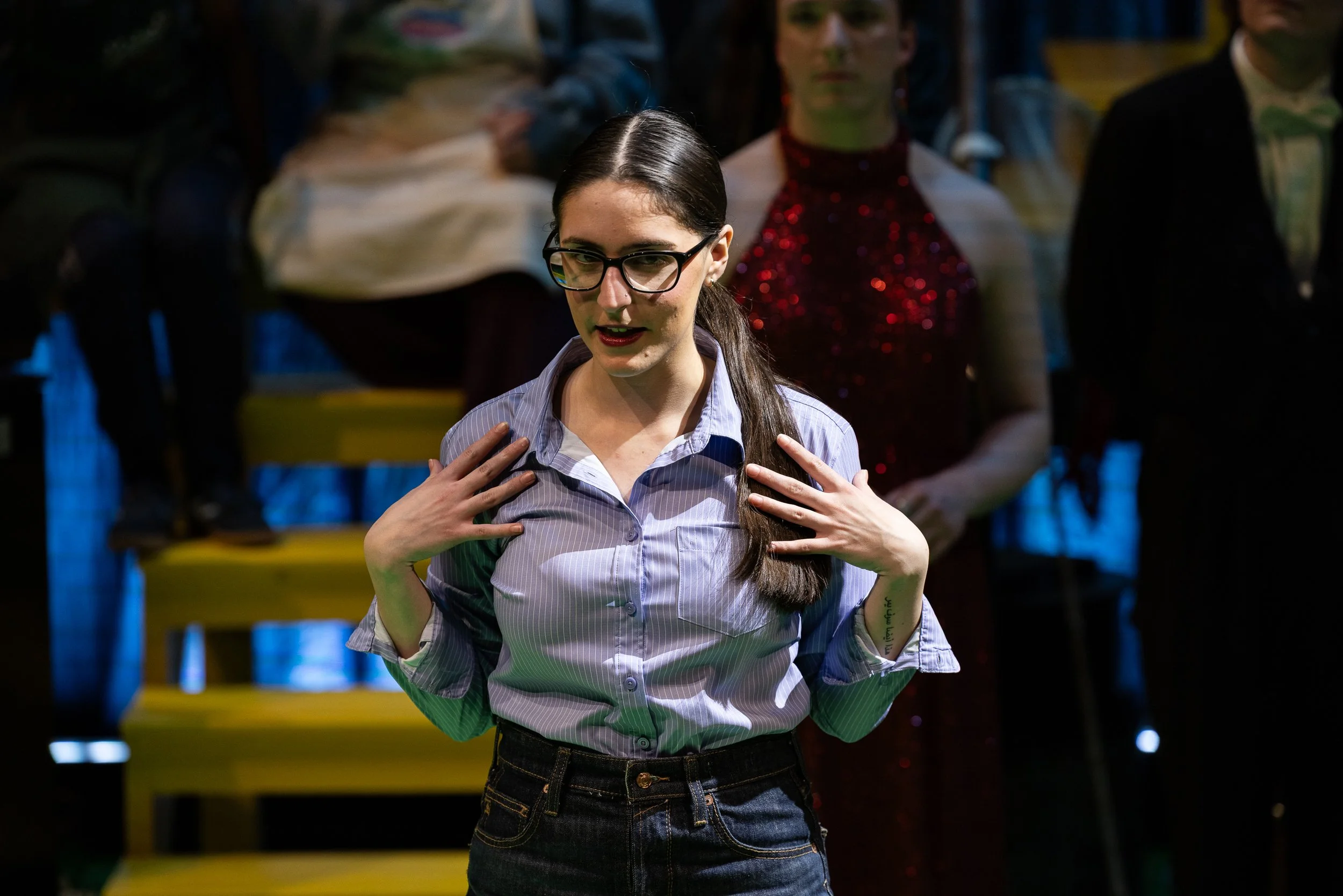 A woman with long dark hair, glasses, and a striped shirt performing on stage with her hands on her chest, surrounded by an audience.