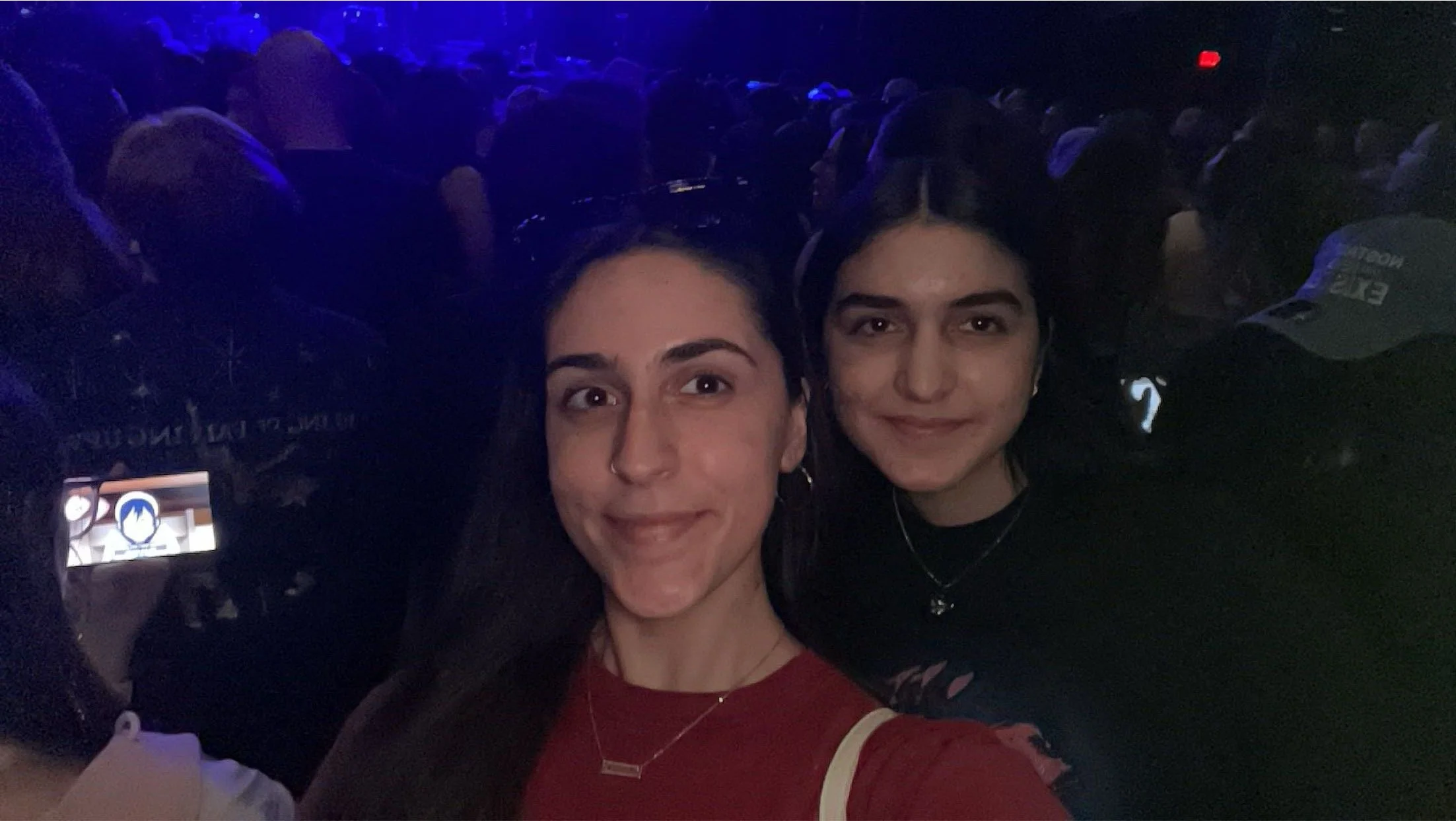 Two young women smiling, taking a selfie at an indoor event or venue with dark lighting and a crowd in the background.