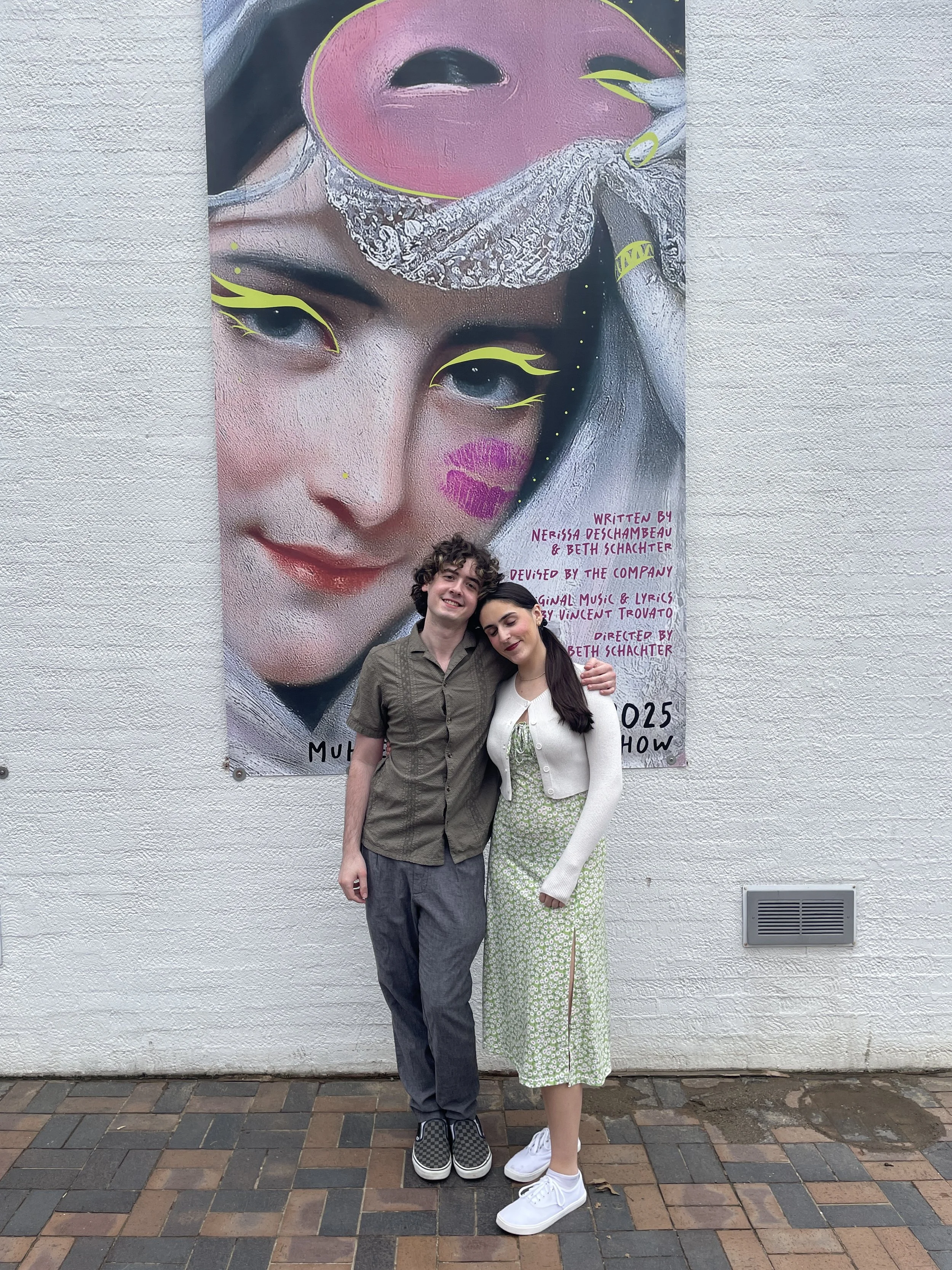 Two young people, a male and a female, standing in front of a large mural of a woman's face. They are smiling and leaning on each other, with the mural artist's name and show information written on it.