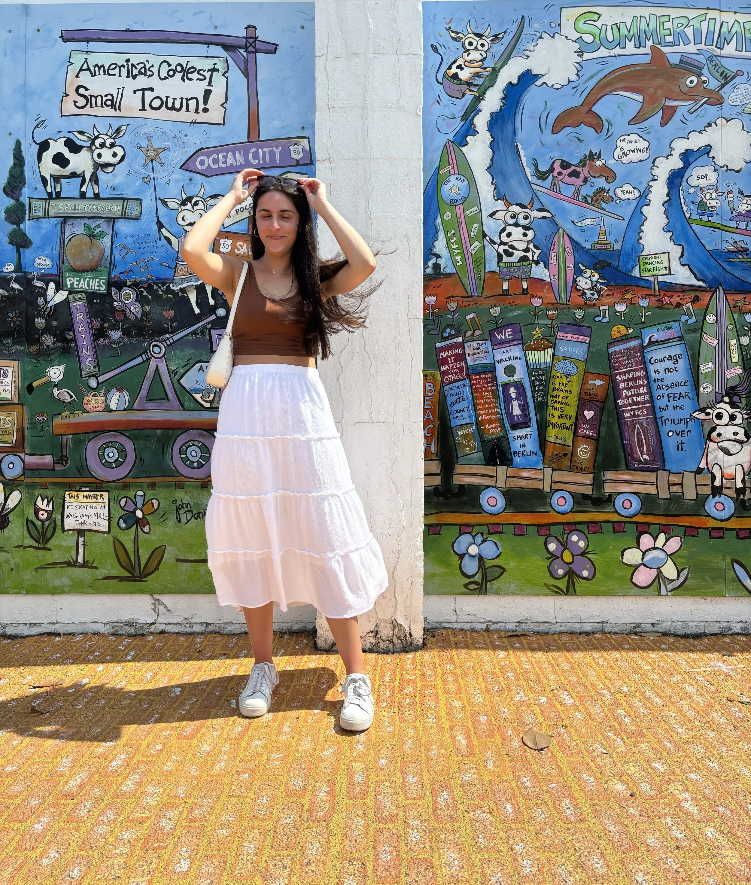 Young woman standing in front of a colorful mural, wearing a brown sleeveless top, a white flowing skirt, white sneakers, and sunglasses, with her eyes closed and hair blowing in the wind.
