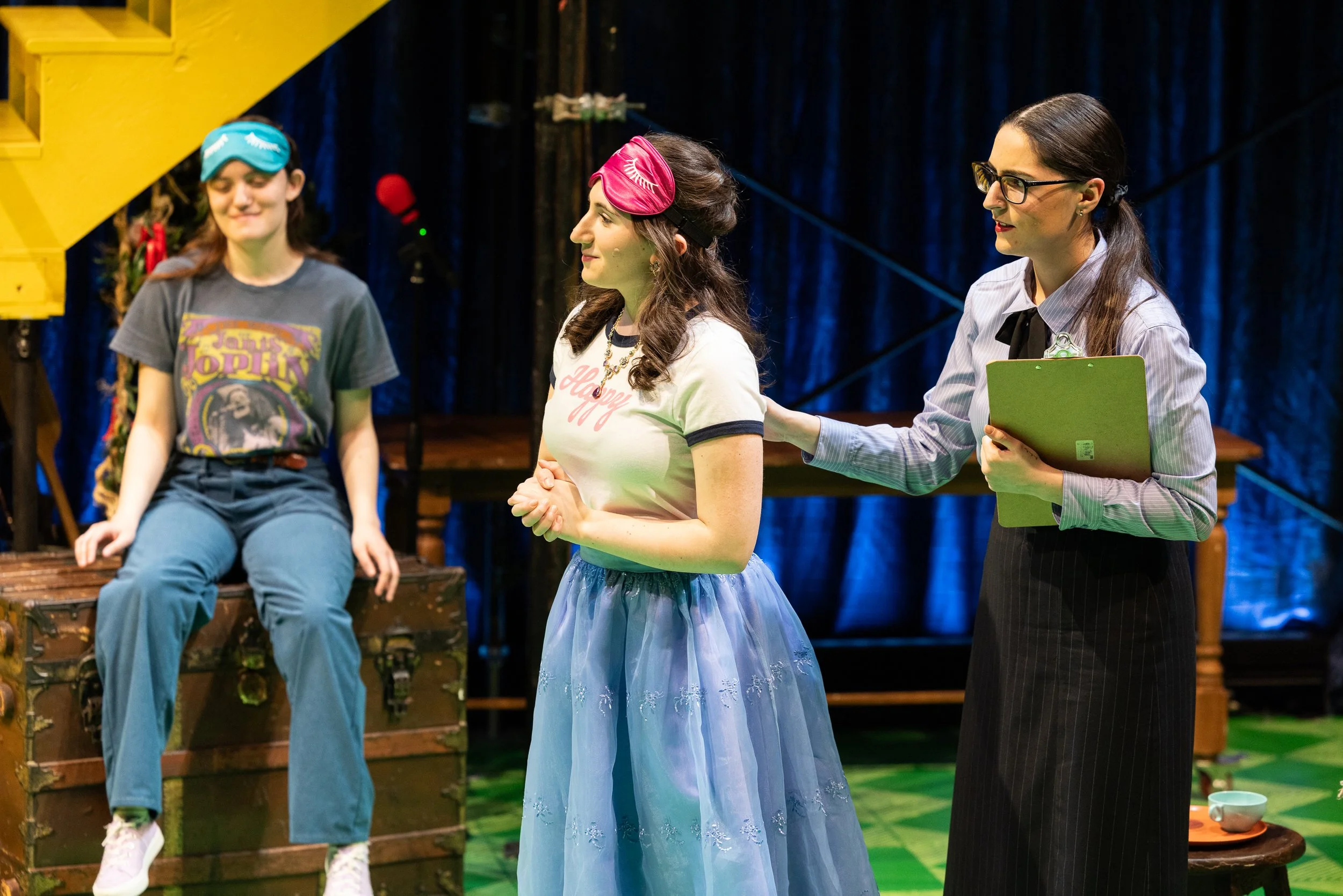 Three women standing on a stage, with one woman in a blue sheer skirt and pink sleep mask, and the other two women in casual clothing and glasses.