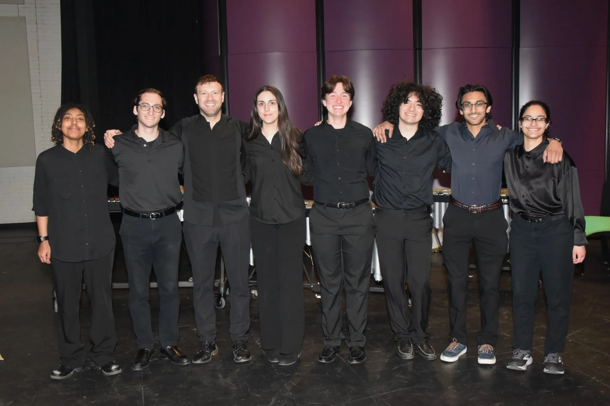 Group of eight diverse people standing closely together with arms around each other, dressed in black, smiling at the camera in a dark indoor setting.