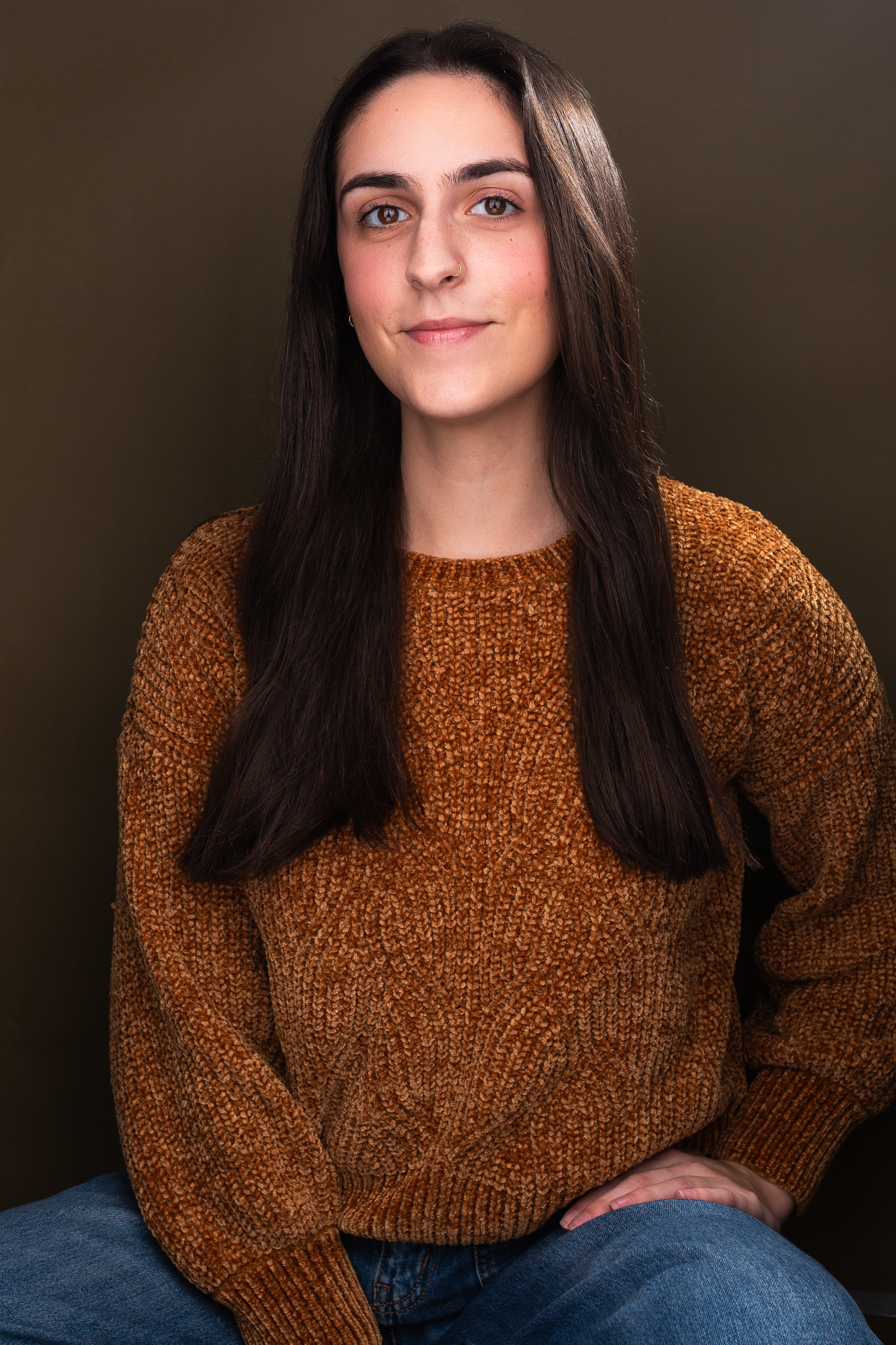 A woman with long dark hair wearing a textured brown sweater and blue jeans, sitting with her hands folded on her lap, against a neutral background.