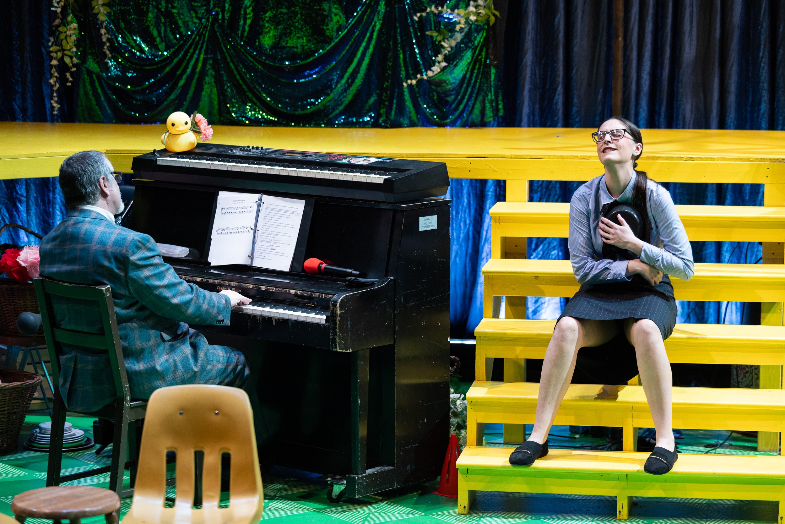 A scene from a stage performance with a woman wearing glasses, a striped shirt, and a skirt sitting on yellow wooden stairs, singing or acting emotionally with her hand on her chest. At the left, a man in glasses and a plaid jacket is playing a black