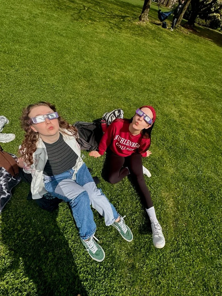 Two girls sitting on grass outdoors, wearing eclipse glasses and making puckered faces. One girl has curly hair, striped shirt, and jeans. The other girl has straight hair, a red hoodie, and black leggings. There are backpacks and trees in the backgr