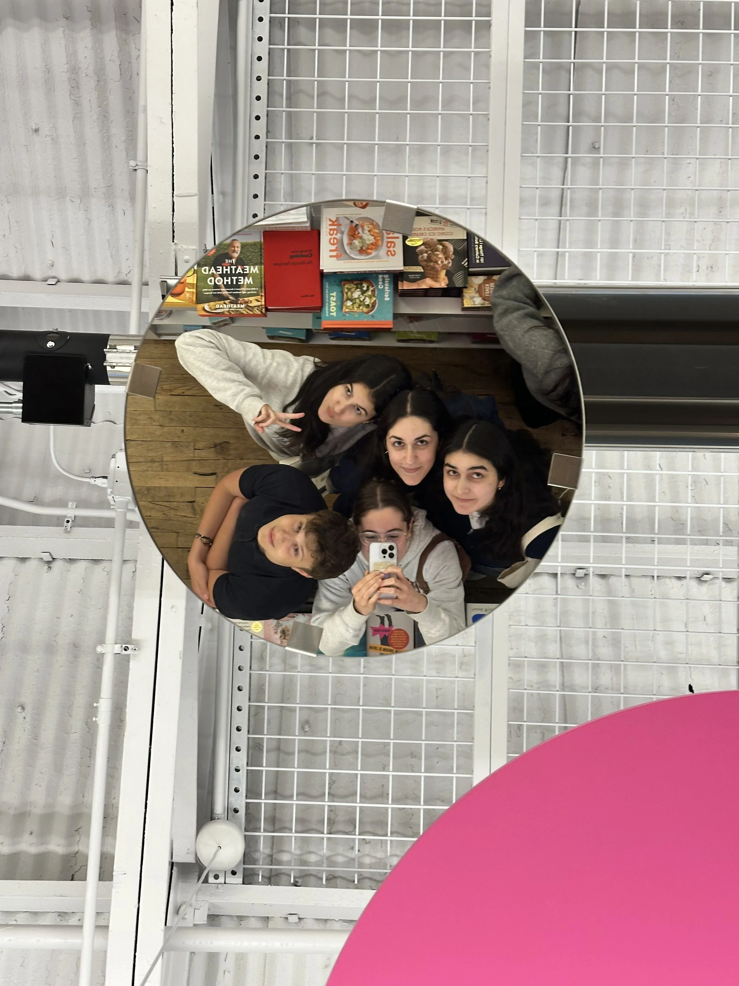 Four young women taking a selfie in front of a mirror at a bookstore, with shelves of books behind them, and a pink table in the foreground.