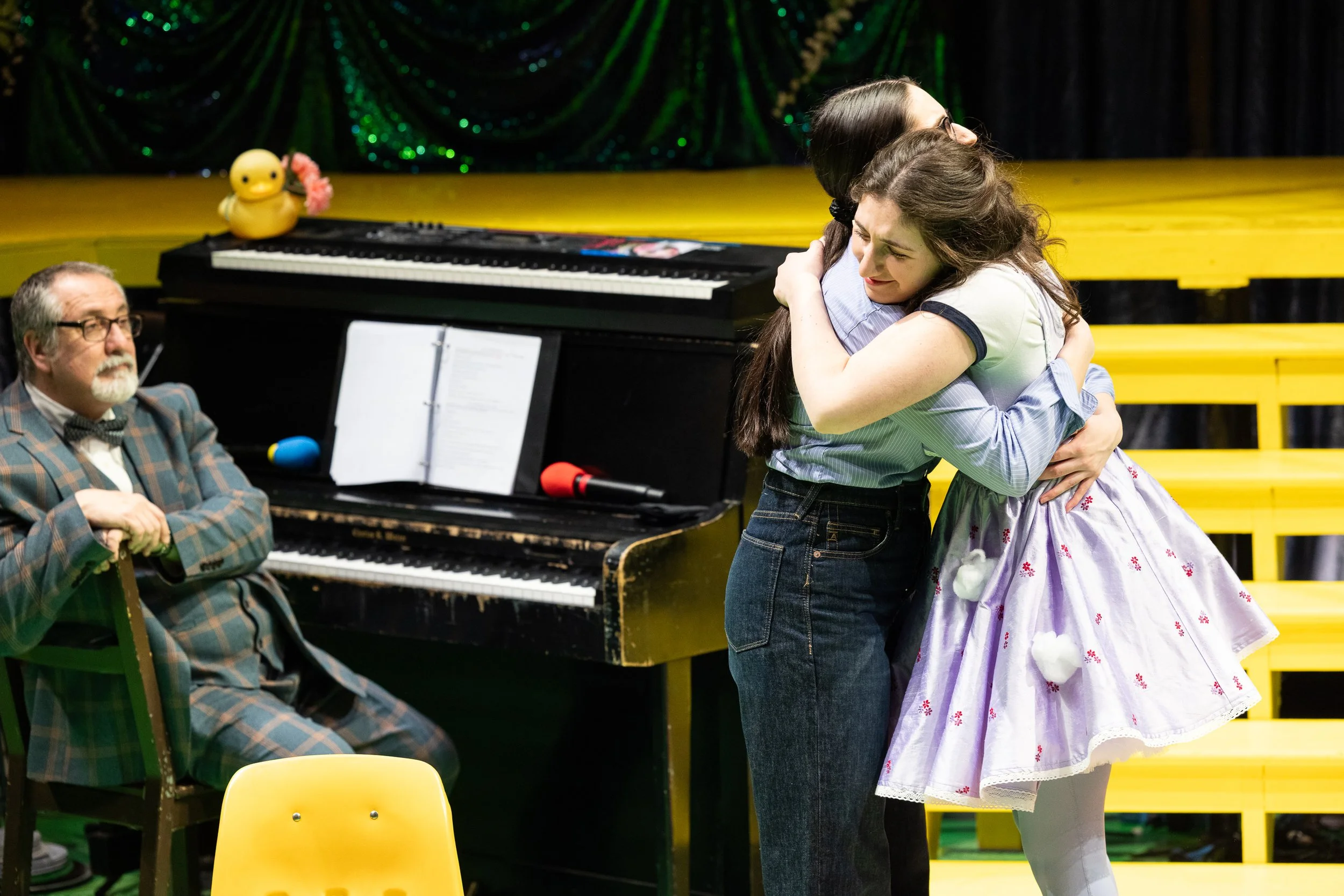 Two girls hugging each other on stage, with a piano and a rubber duck toy in the background, and a man sitting nearby observing.