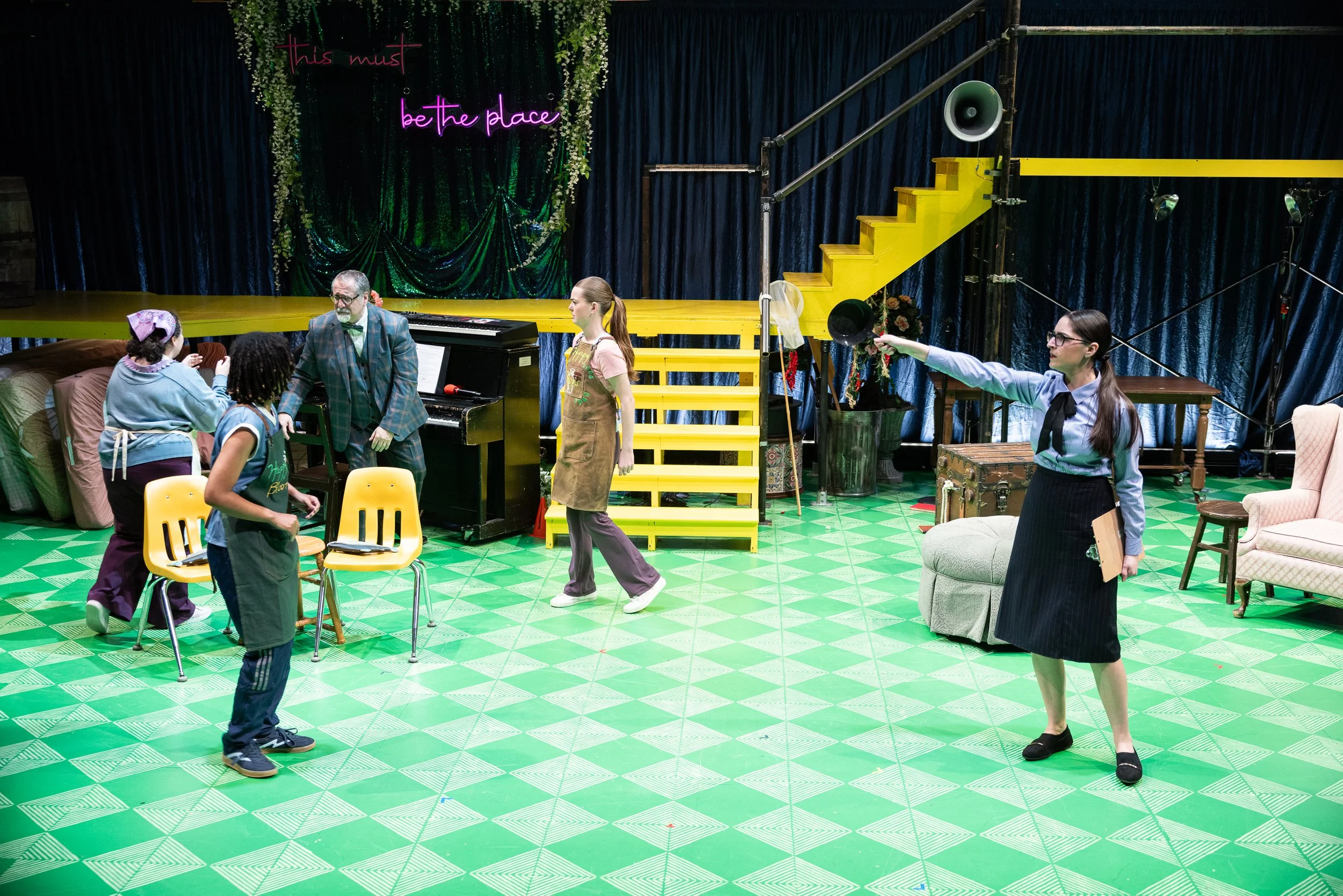 People on stage preparing for a theatrical performance in a colorful setting with a dark green curtain, yellow stairs, and a neon sign that says 'this must be the place.'