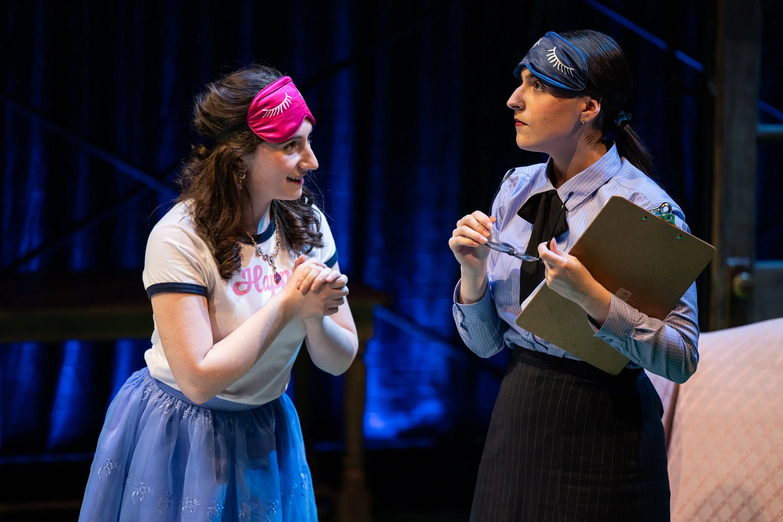 Two women wearing sleep masks with eyelash designs on a stage, engaged in conversation. One wears a pink mask with the word "Happy" and a blue skirt, the other wears a black mask with a white shirt, black tie, and holds glasses and a clipboard.